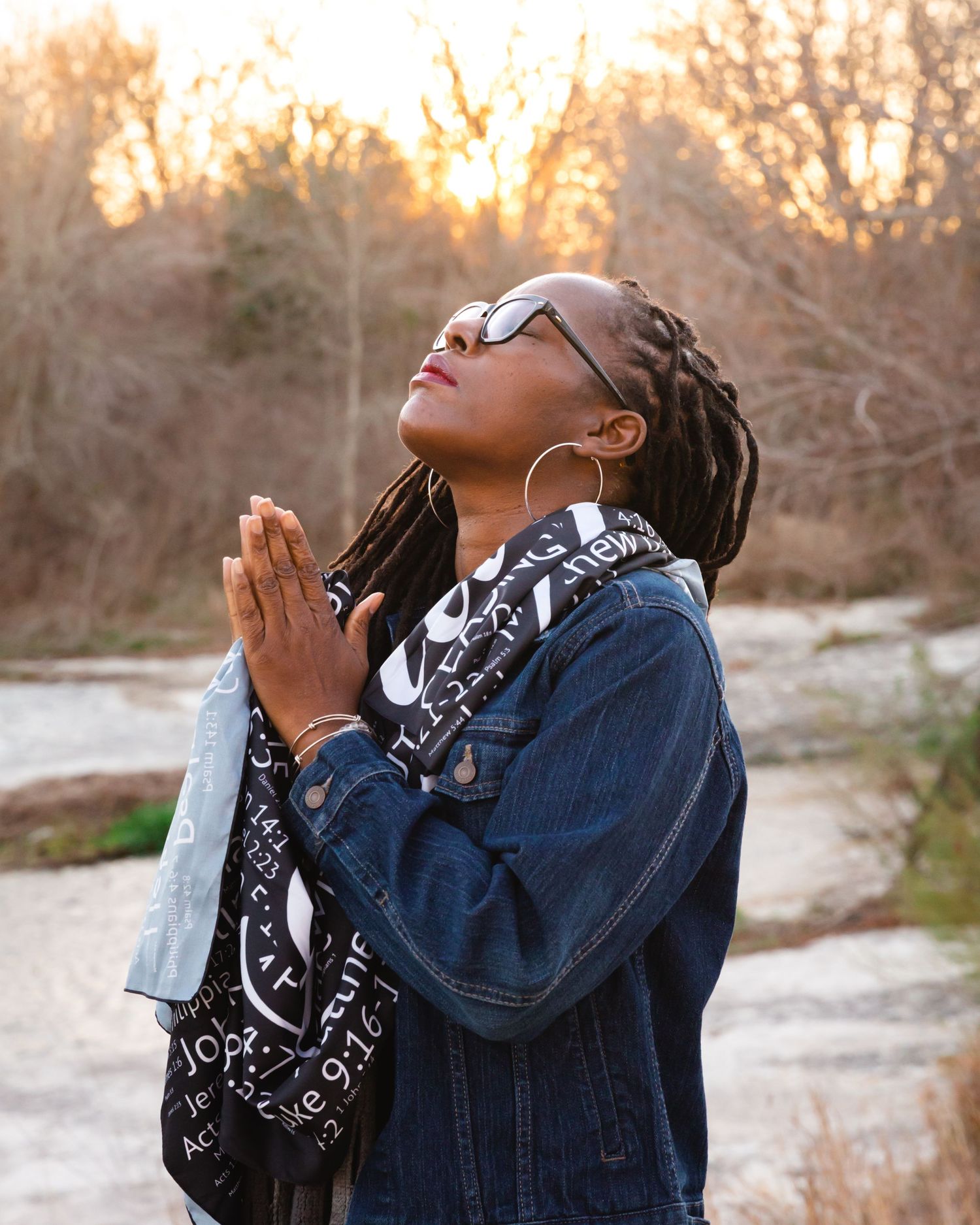 A person in denim jacket and sunglasses prays outdoors during golden sunset.