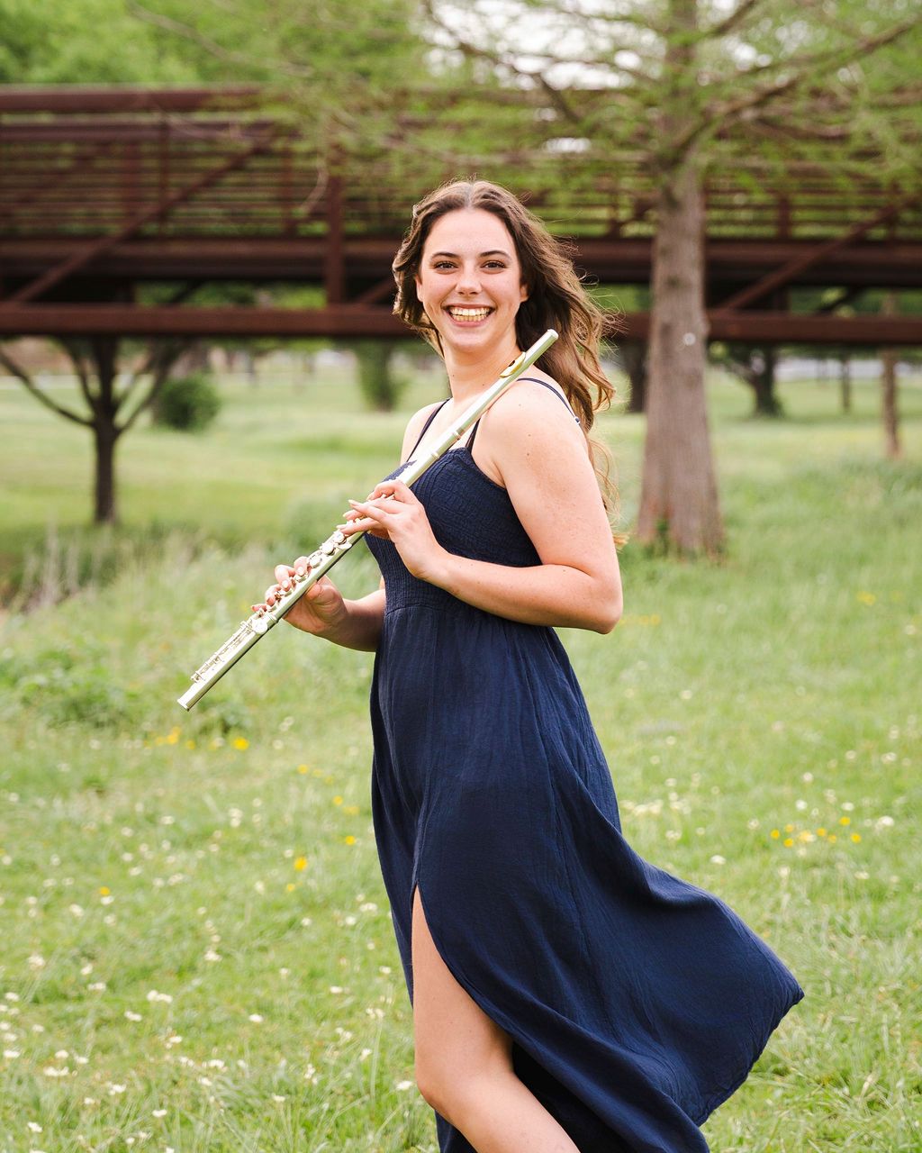 A musician in a flowing navy dress poses with her flute in a sunny park setting.