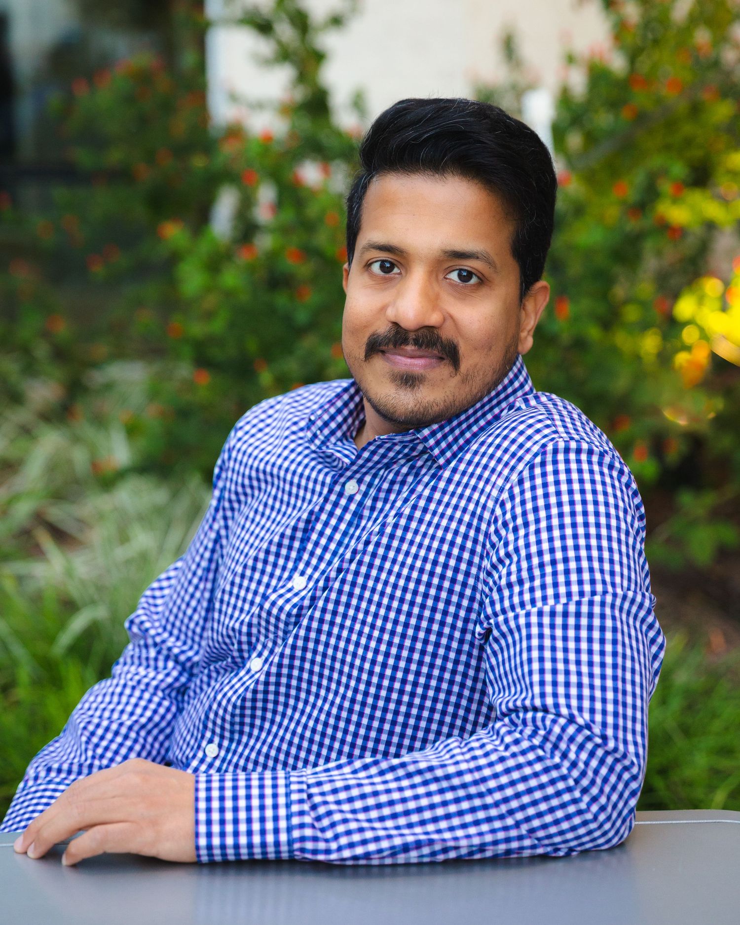 Professional headshot series of person in blue checkered button-down shirt against natural outdoor background.