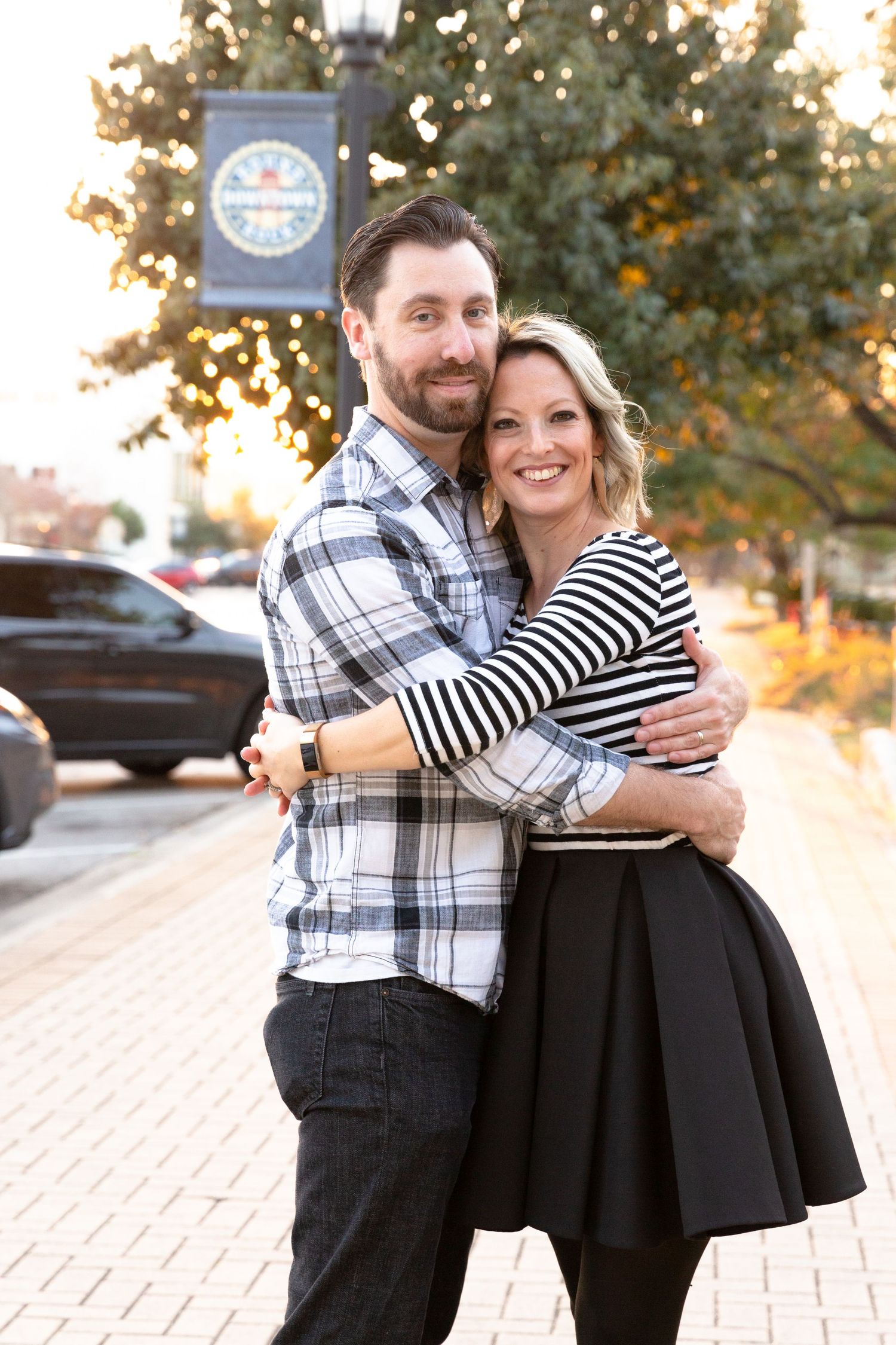 A couple embraces on a brick sidewalk at sunset, wearing plaid and stripes in a casual outdoor setting.