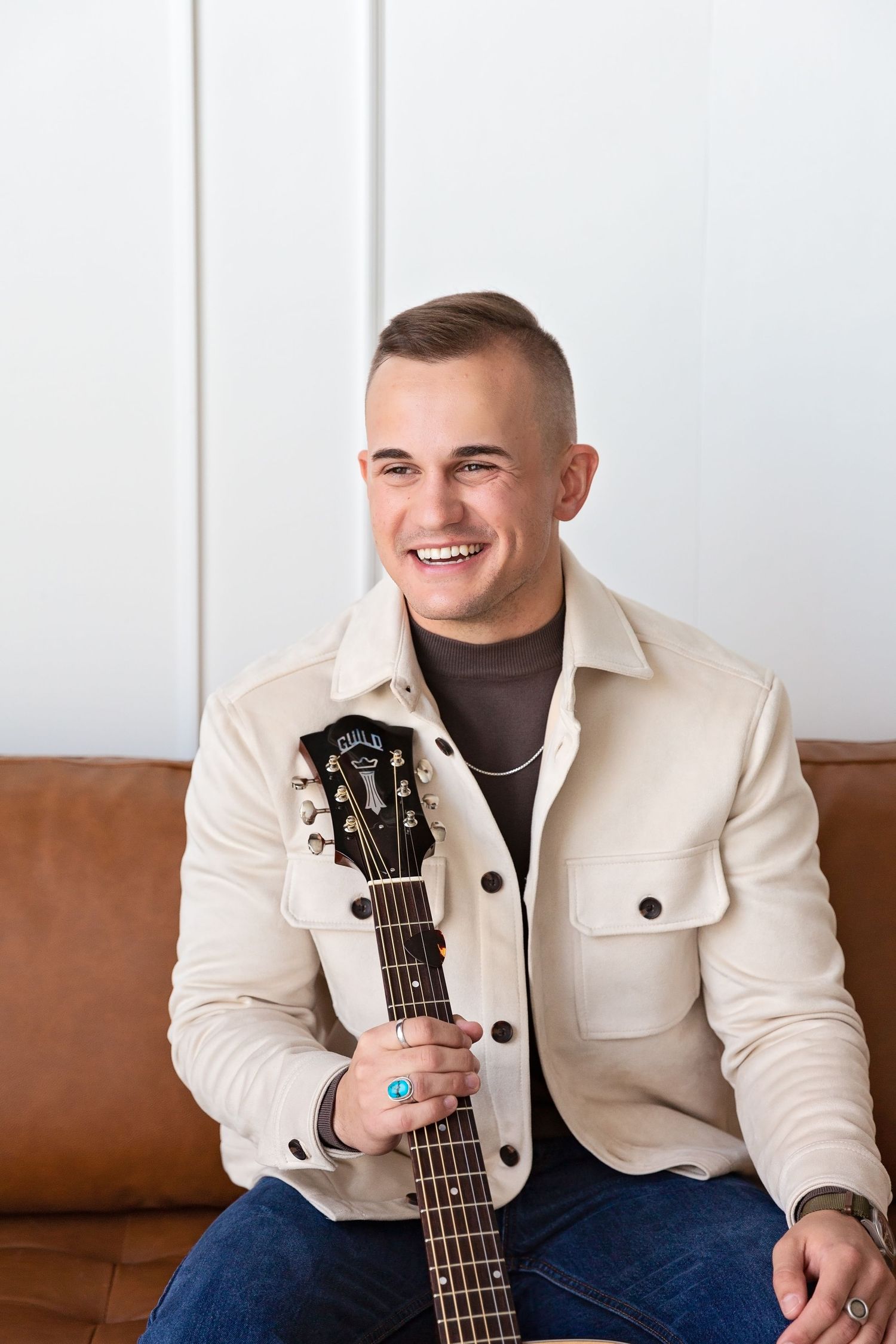 Musician headshot in cream jacket poses with acoustic guitar on brown leather couch. Professional head shot photographer.