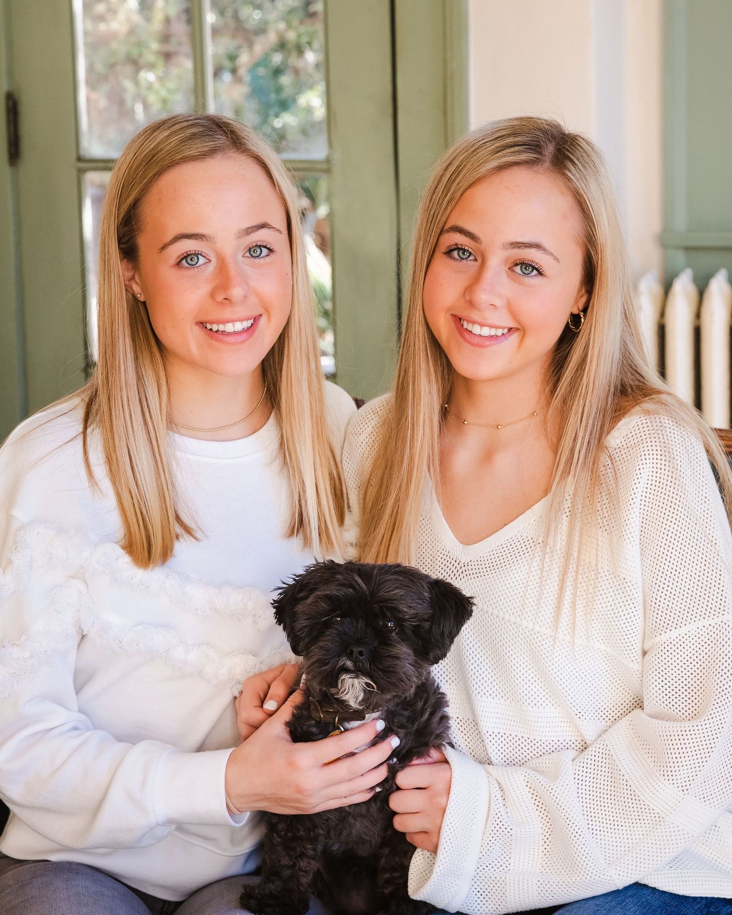 Two women in white sweaters pose with their small black dog in a bright indoor setting.