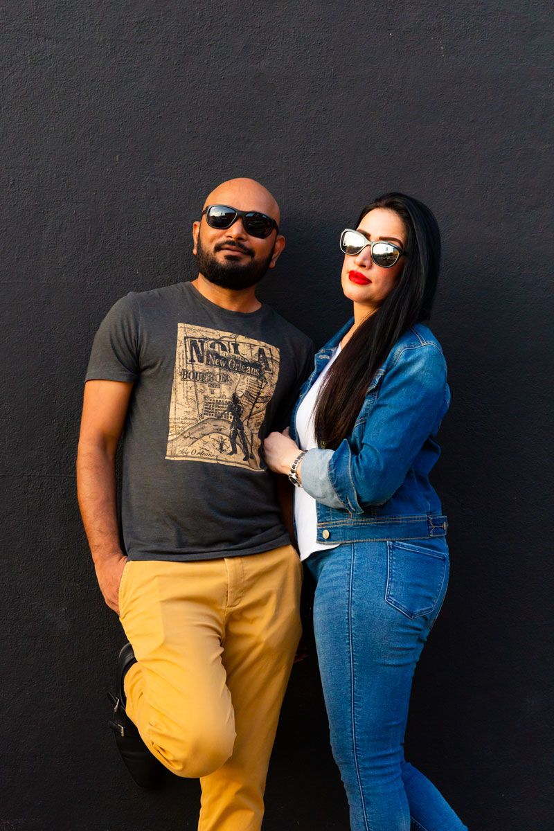 Stylish couple poses against black wall wearing sunglasses and casual attire.