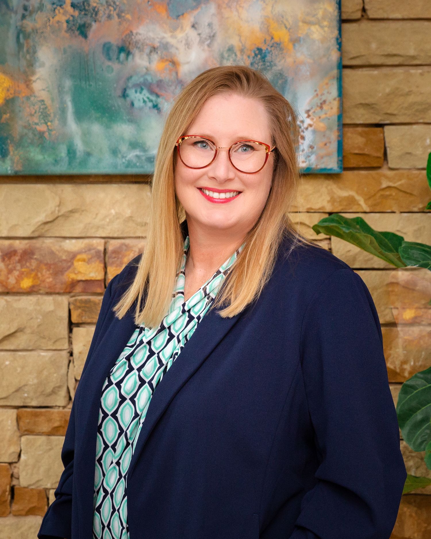 Professional headshot of person in navy blazer and patterned blouse against stone wall backdrop.