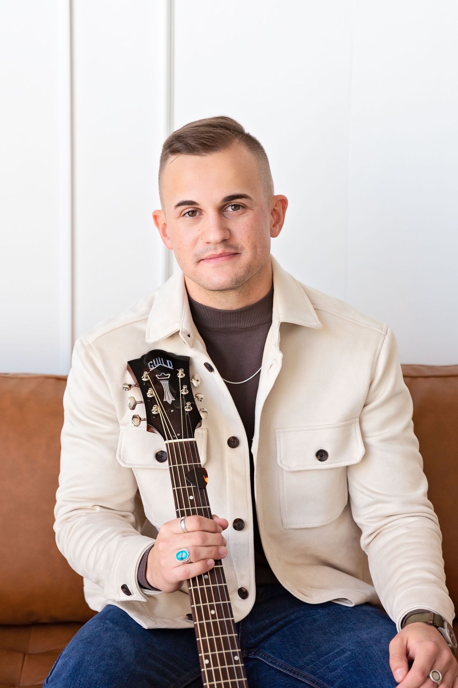 Portrait series of person in cream jacket holding acoustic guitar on brown leather couch.