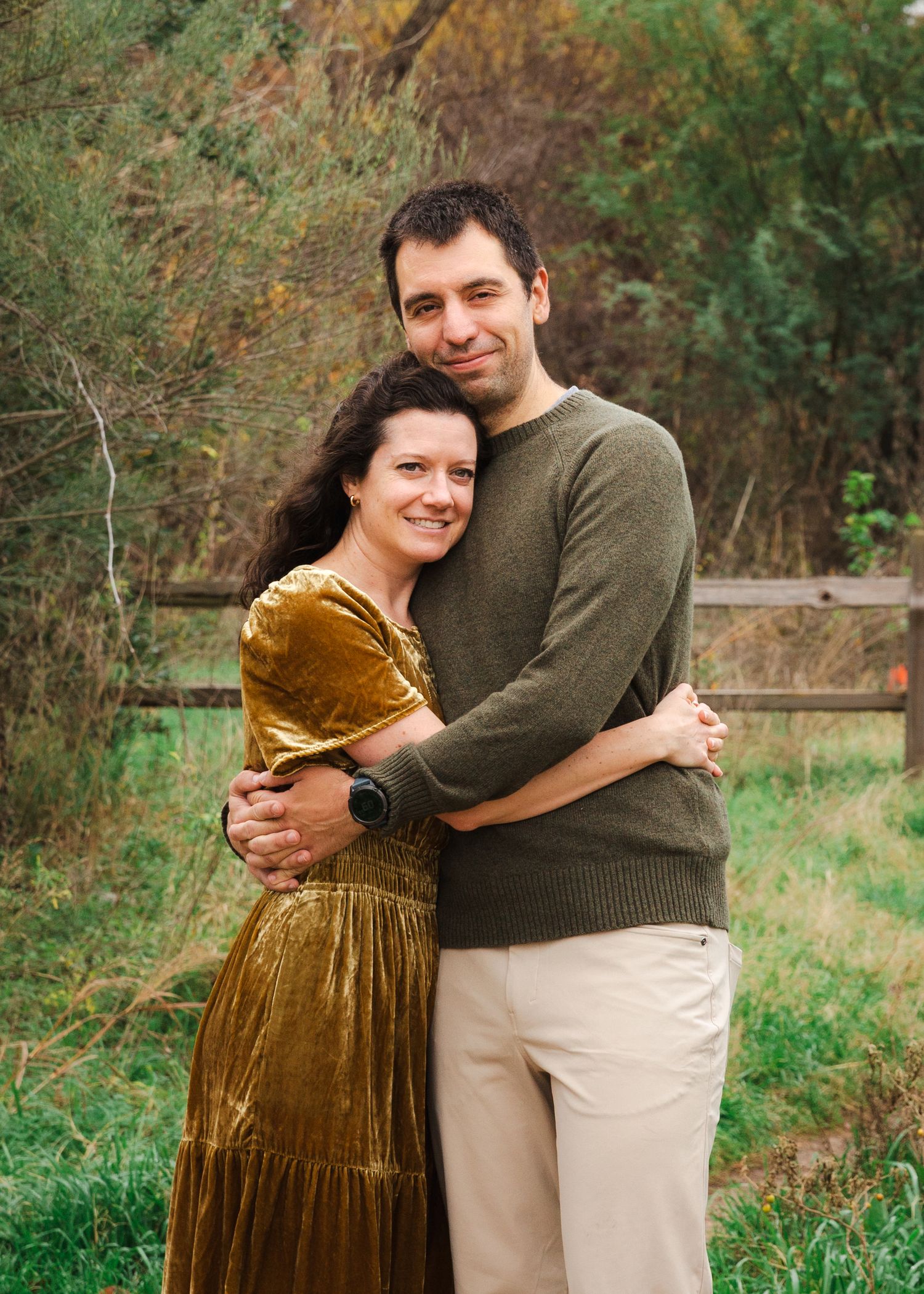 A couple embraces outdoors, with one wearing an olive sweater and the other in a gold velvet dress.