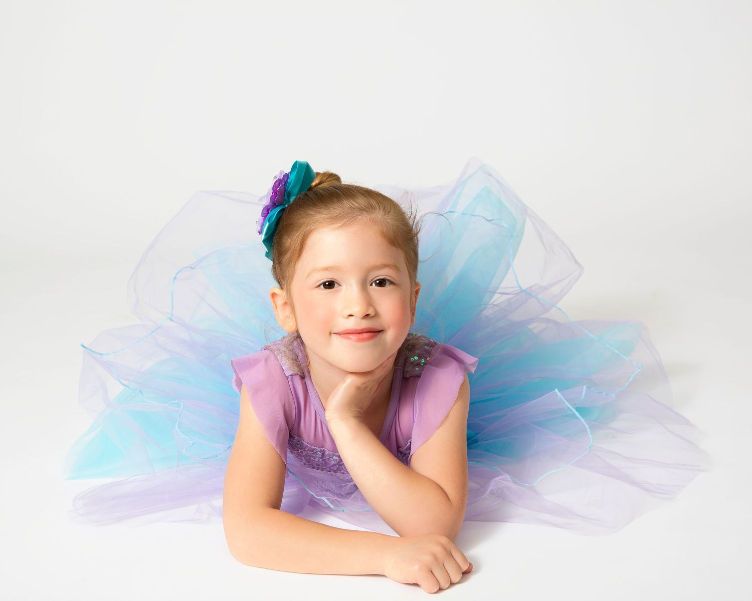Young dancer in purple tulle dress poses gracefully against white background.