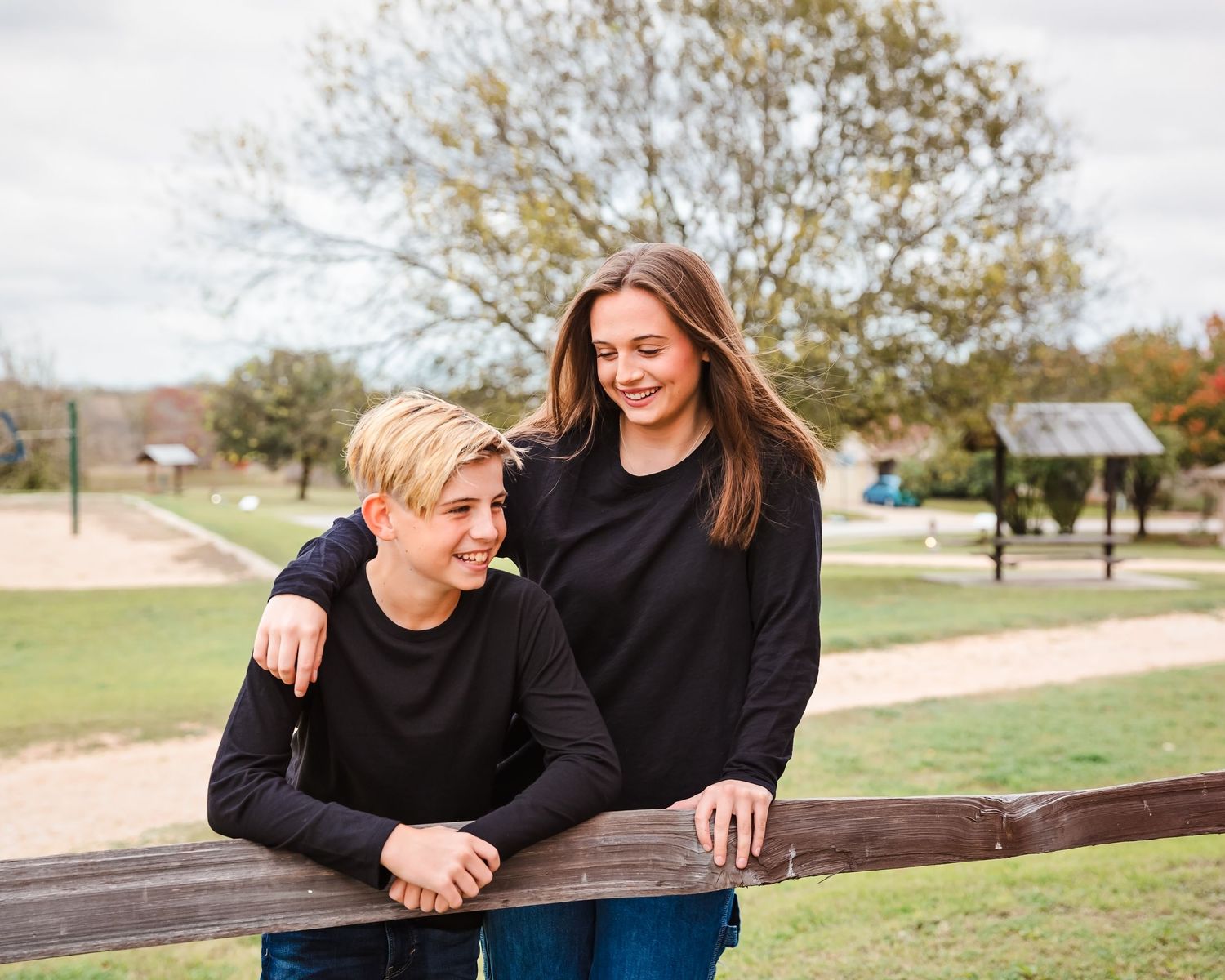 A couple in matching black sweaters sharing a tender moment while leaning on a wooden fence in a park setting.