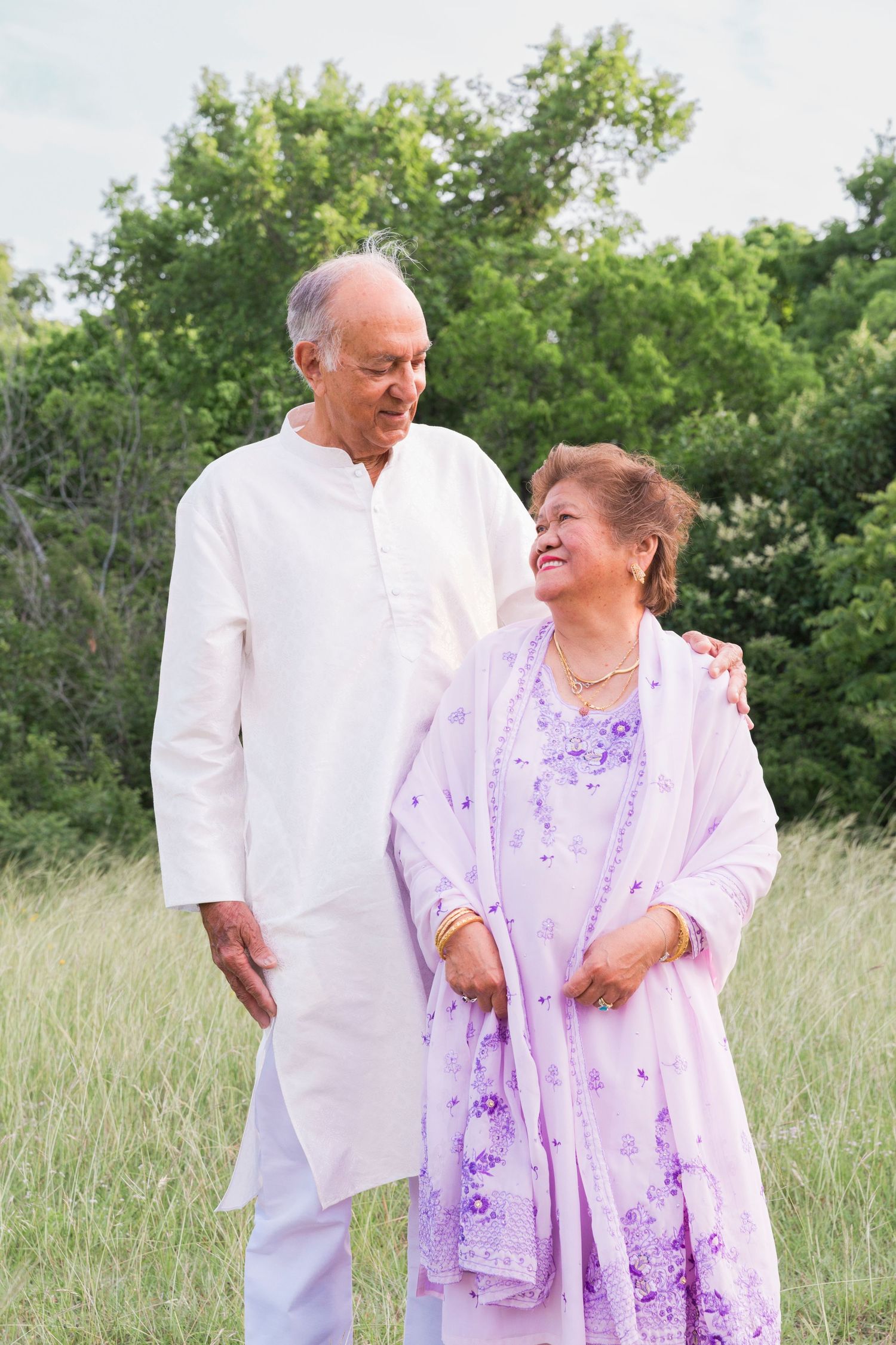 A couple in white and purple traditional attire stands together in a grassy field.
