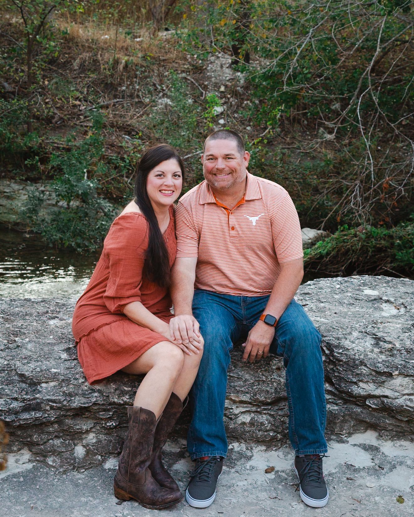 A couple sits together on a rocky outcrop near a wooded area.