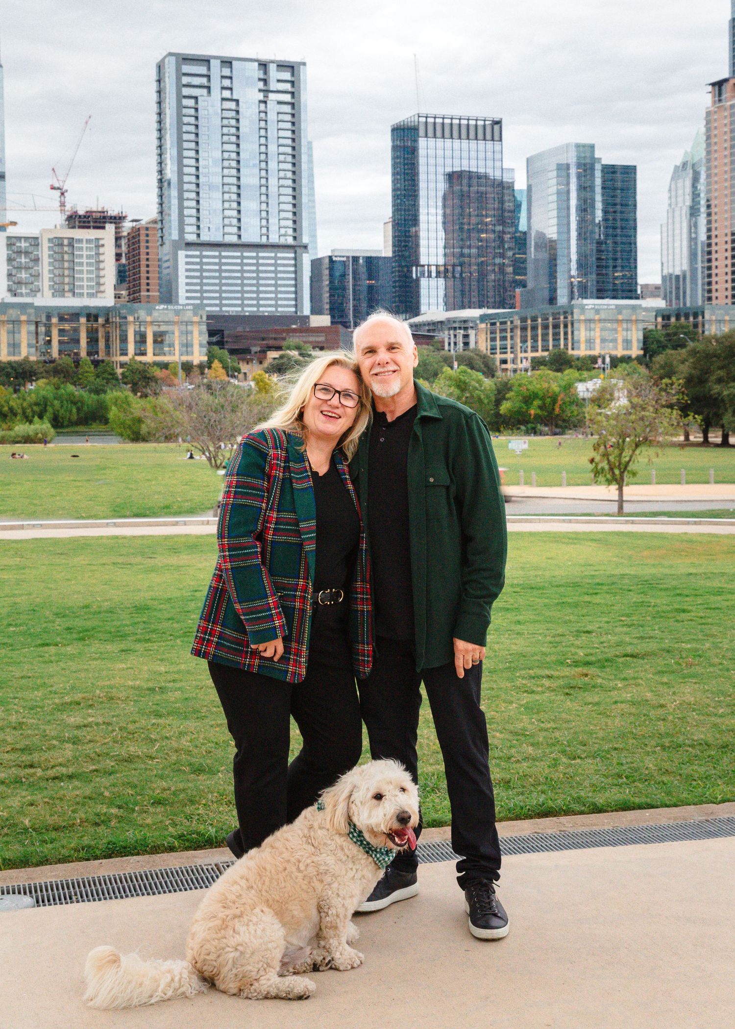 A couple poses with their fluffy white dog against the Austin skyline on a cloudy day.