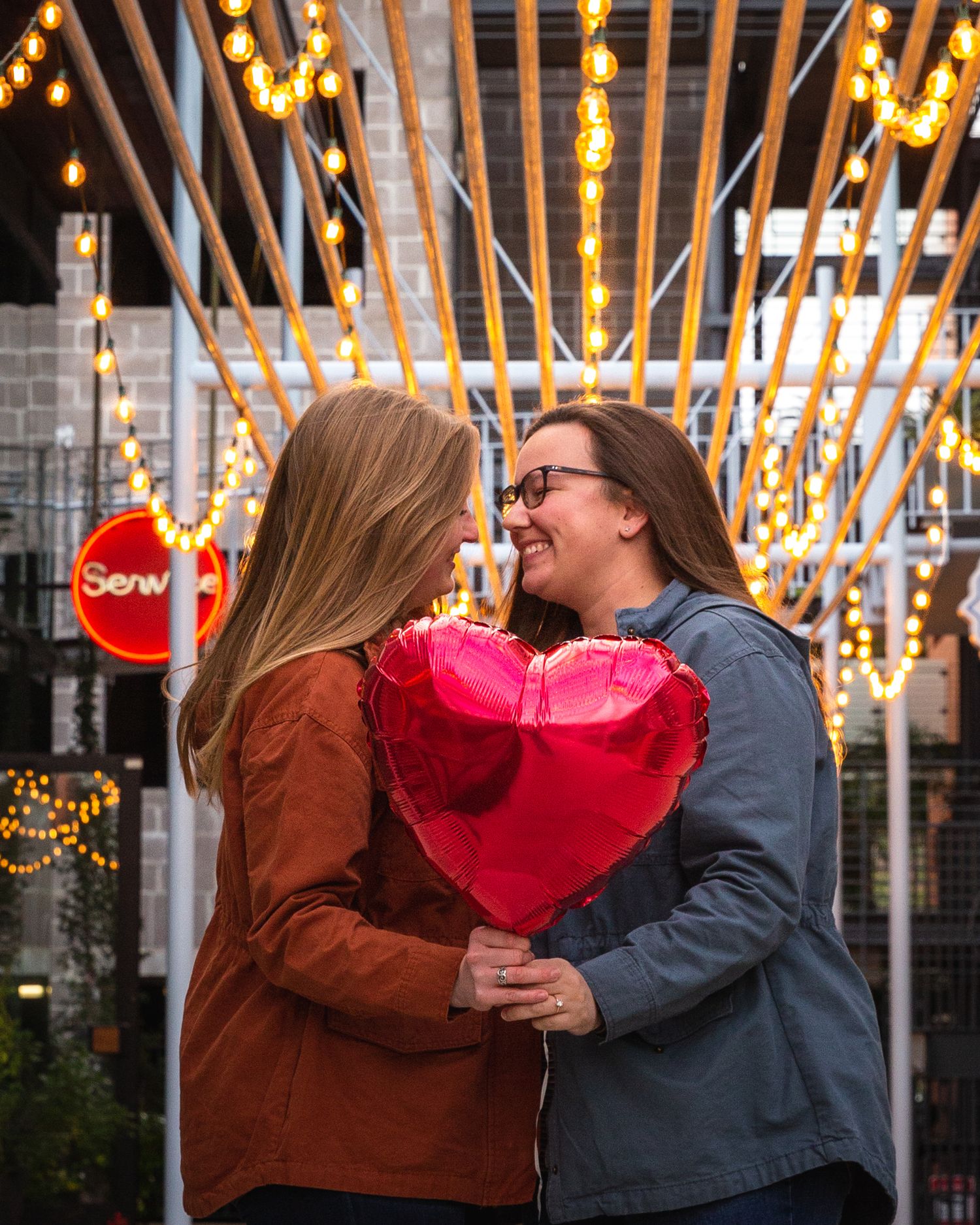 Two people share a romantic moment with a heart balloon under string lights.