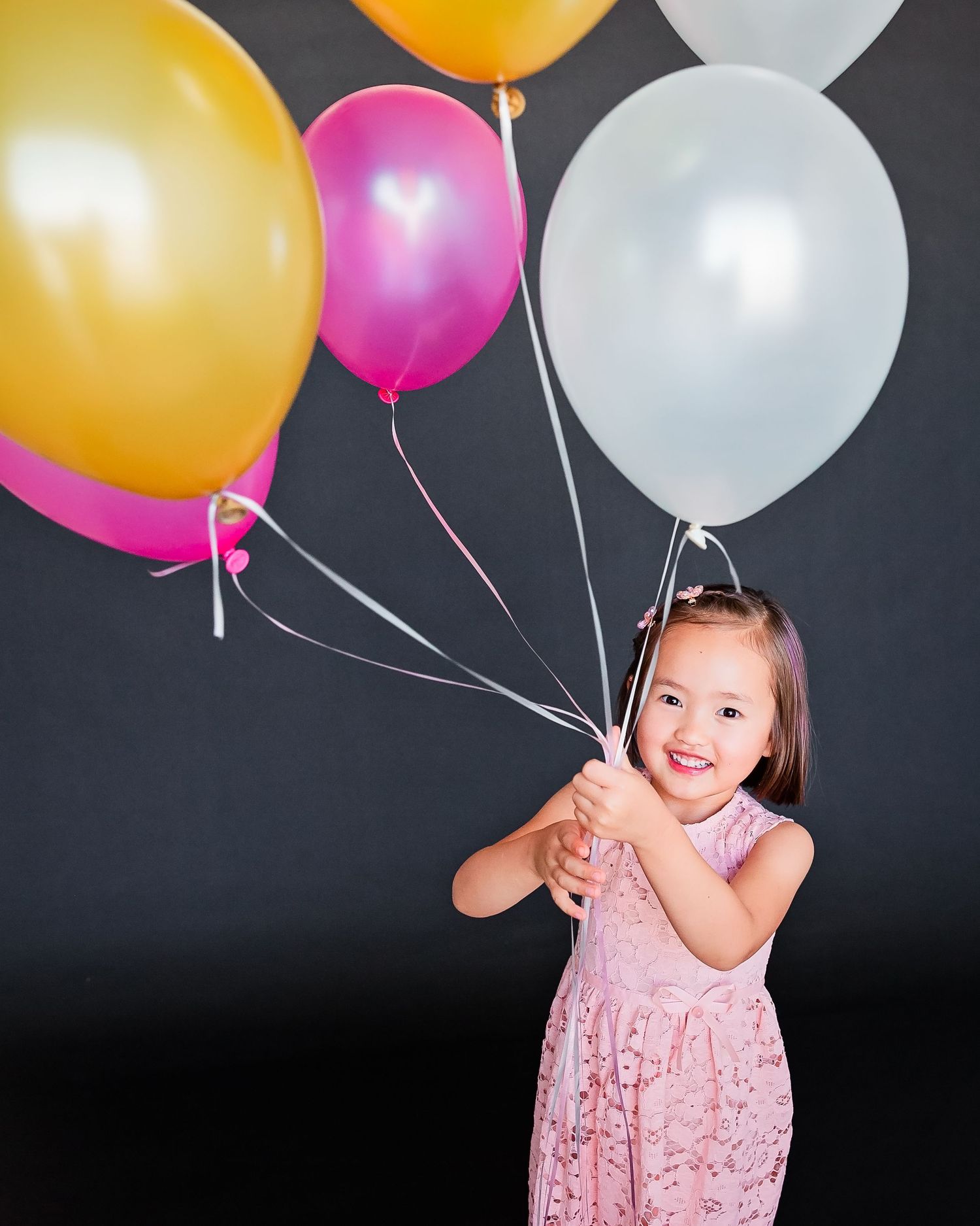 Young child in pink dress holding colorful balloons against dark background.