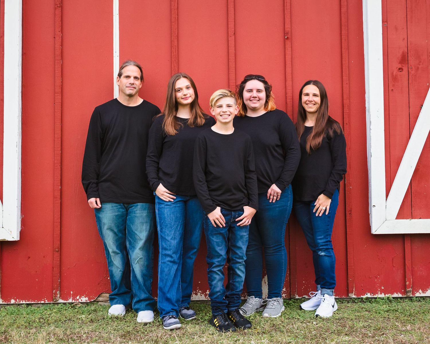 A family wearing coordinated black tops and blue jeans poses together against a bright red barn wall.