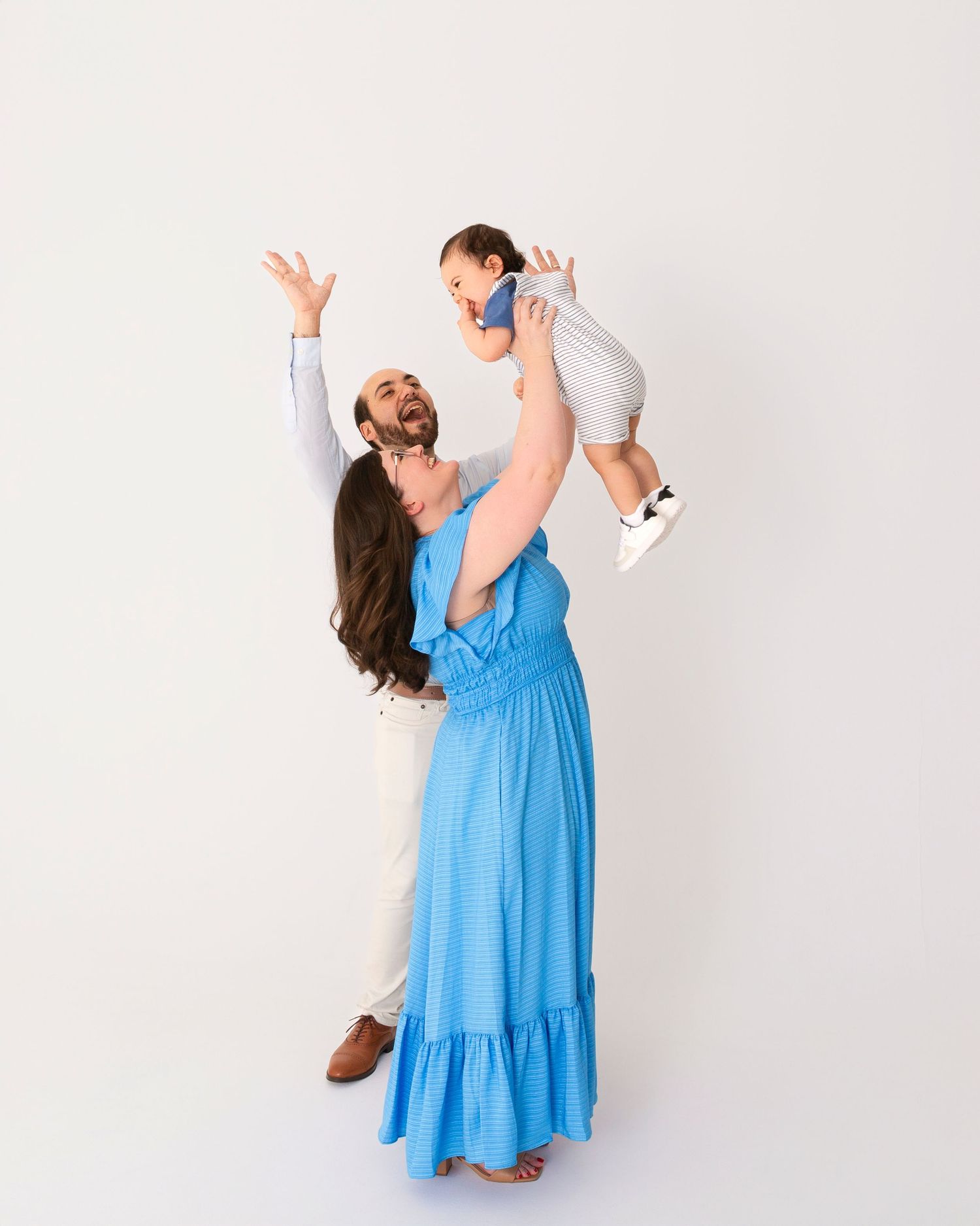 A woman in a flowing blue maxi dress lifts a small child joyfully against a white background.