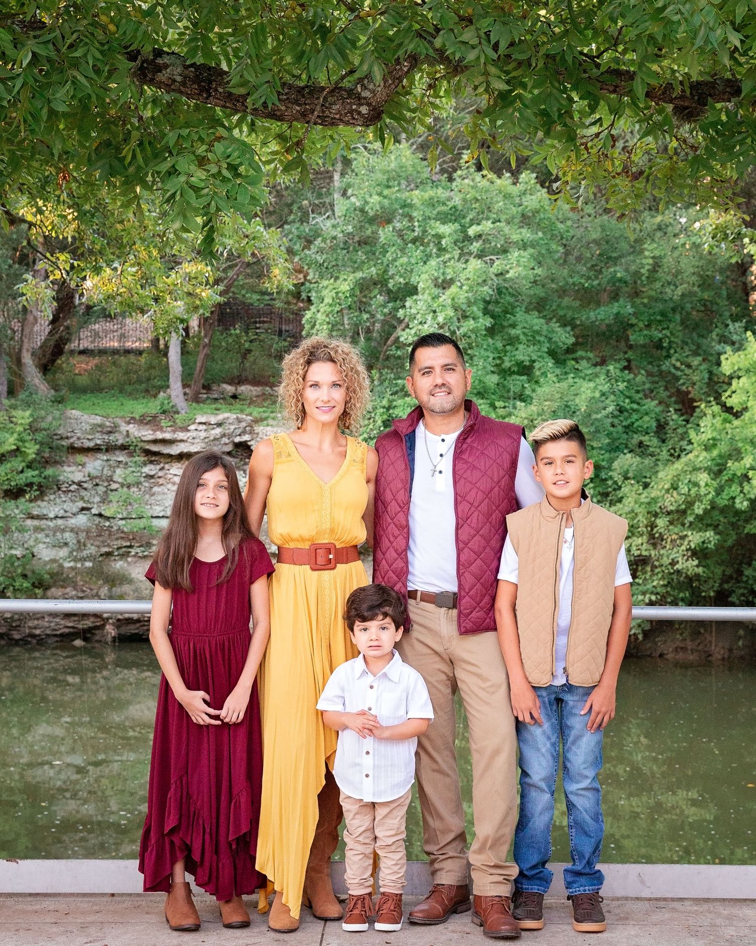 A family wearing coordinated casual outfits poses together by a pond in a natural outdoor setting.