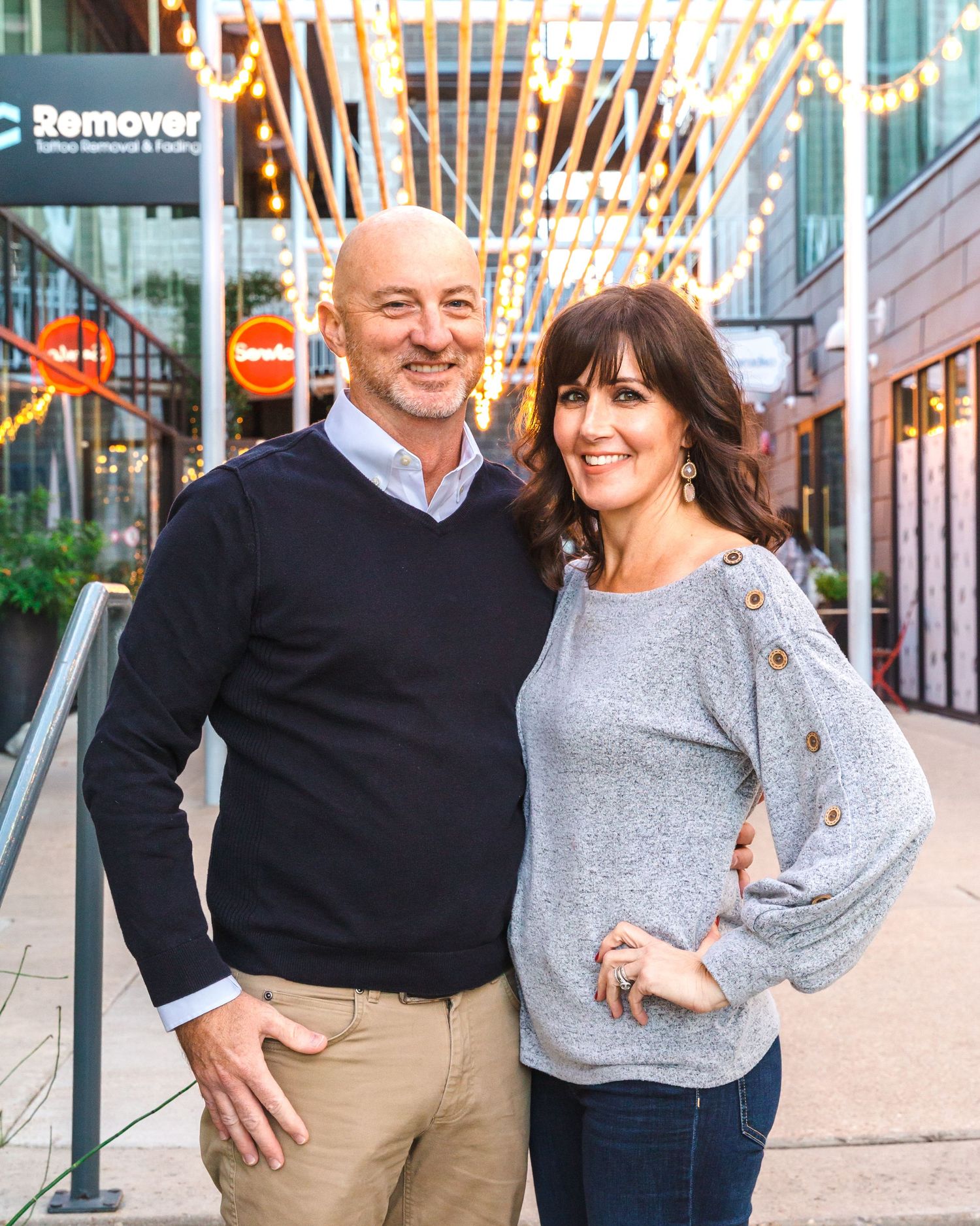 A couple poses in casual winter wear outside an urban shopping center with decorative lighting.