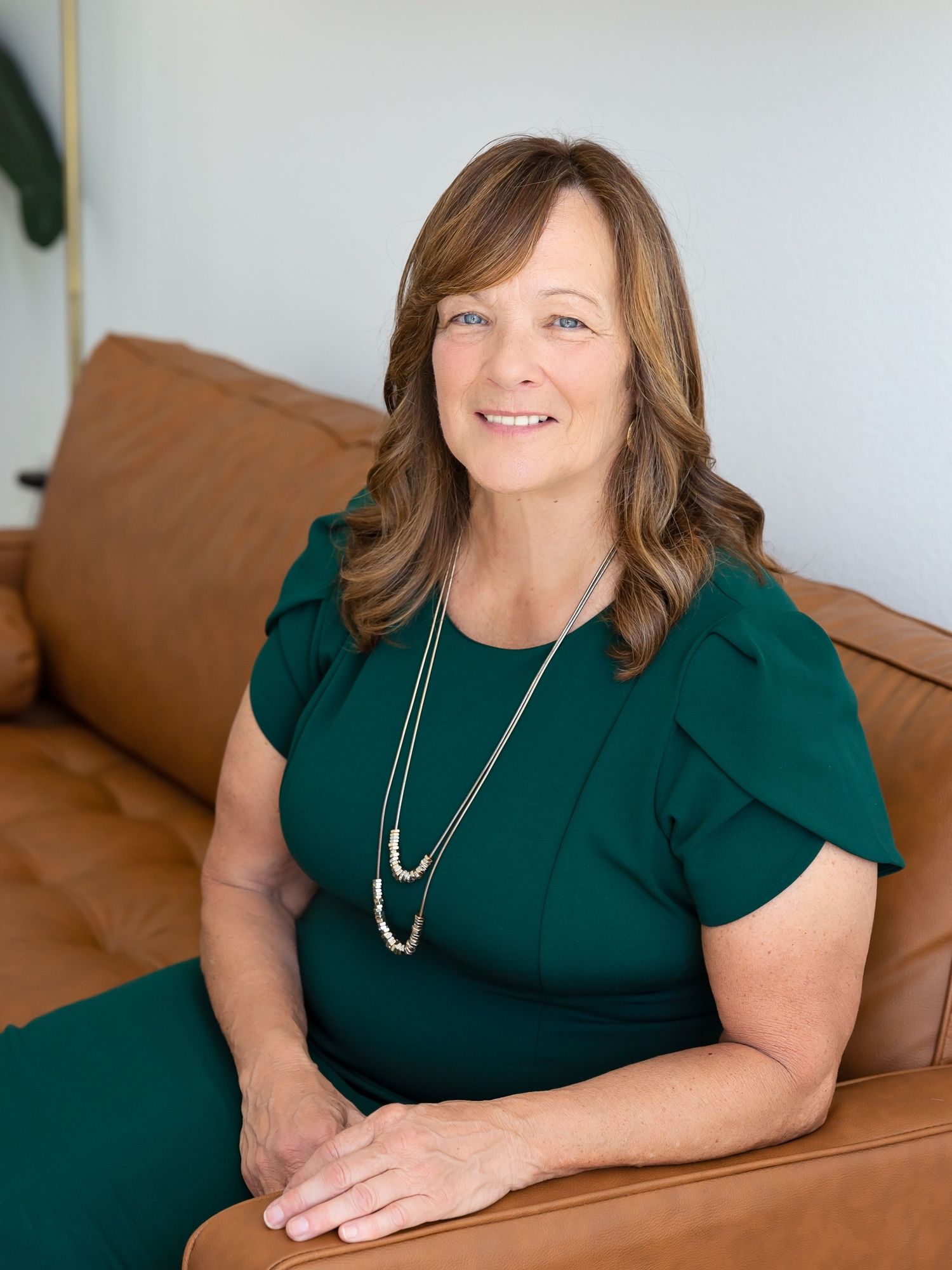 Professional portraits in emerald green top with necklace on brown leather couch.