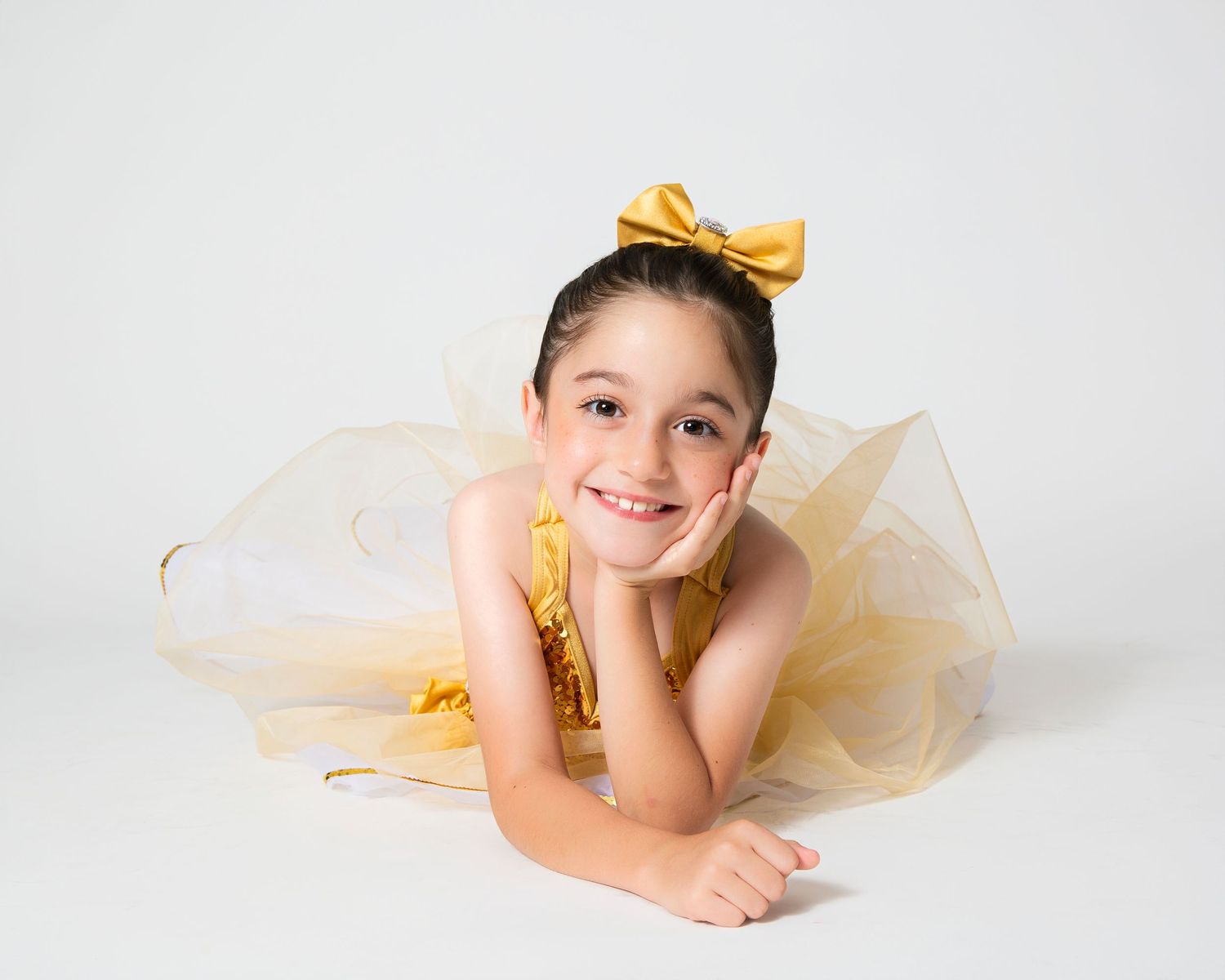 Dancer in gold tulle dress lying elegantly on white studio floor.