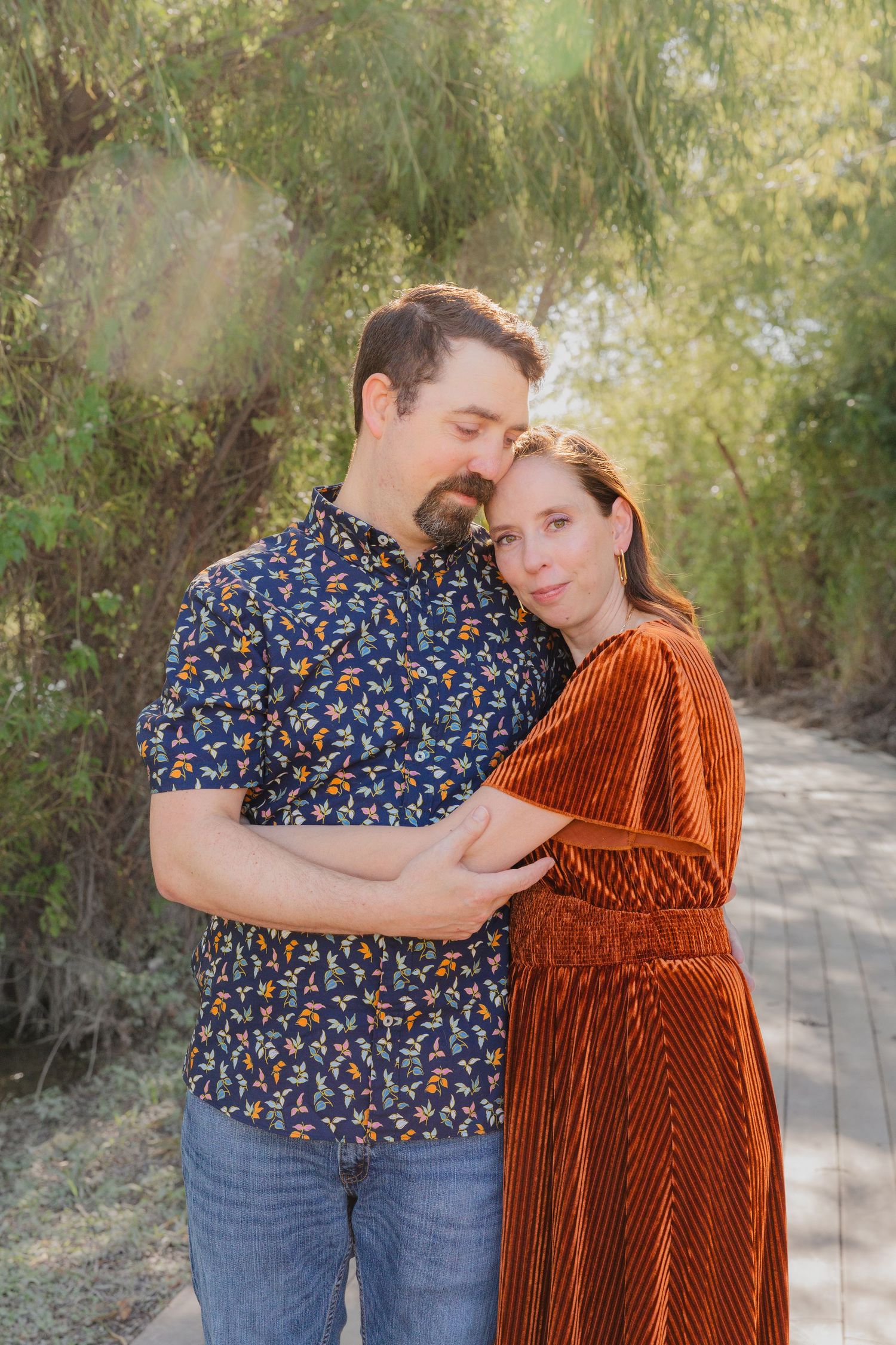A couple embraces outdoors, with one wearing a floral shirt and the other in a rust-colored velvet dress.