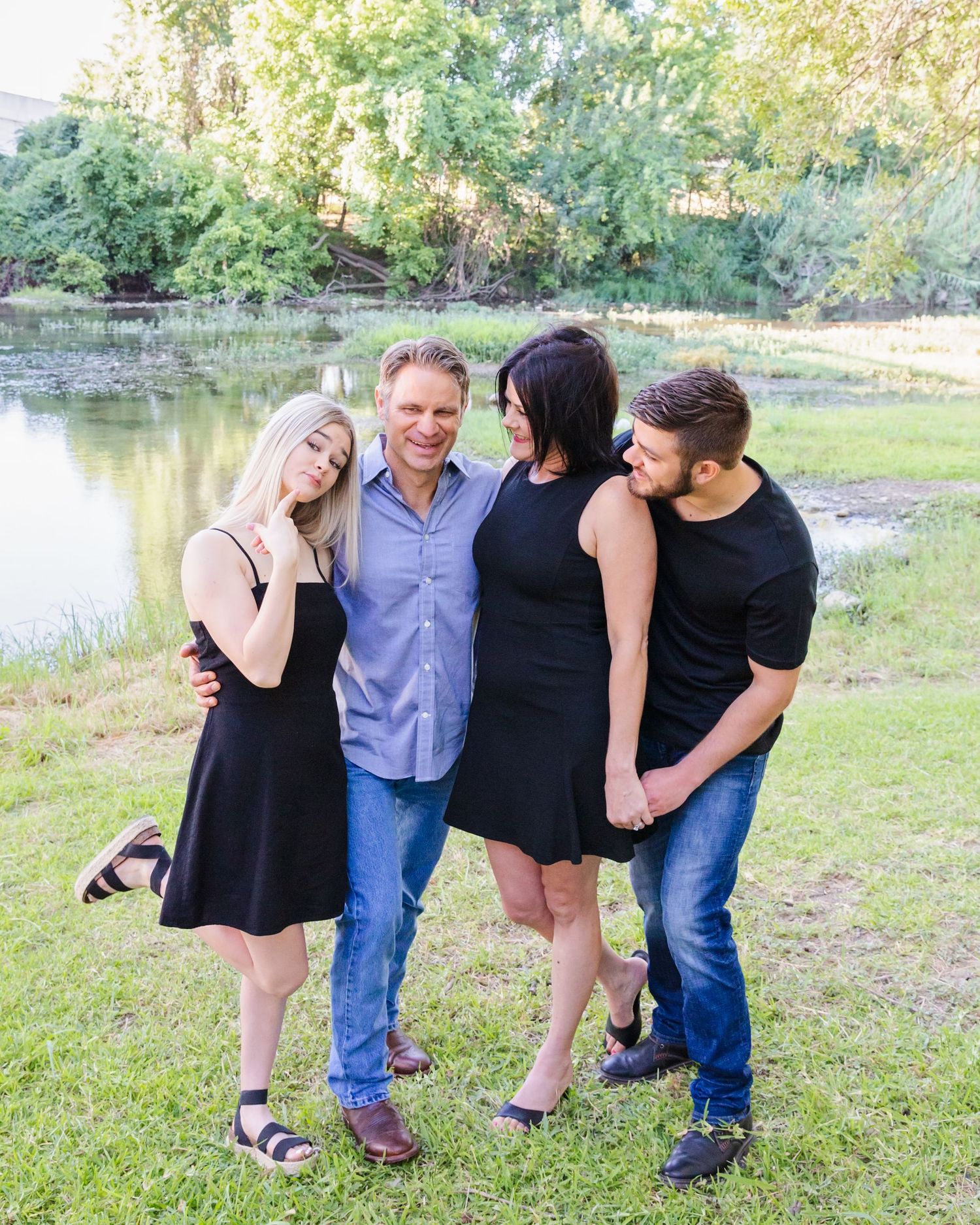 Four people laughing together while posing playfully in an outdoor setting with a pond in the background.