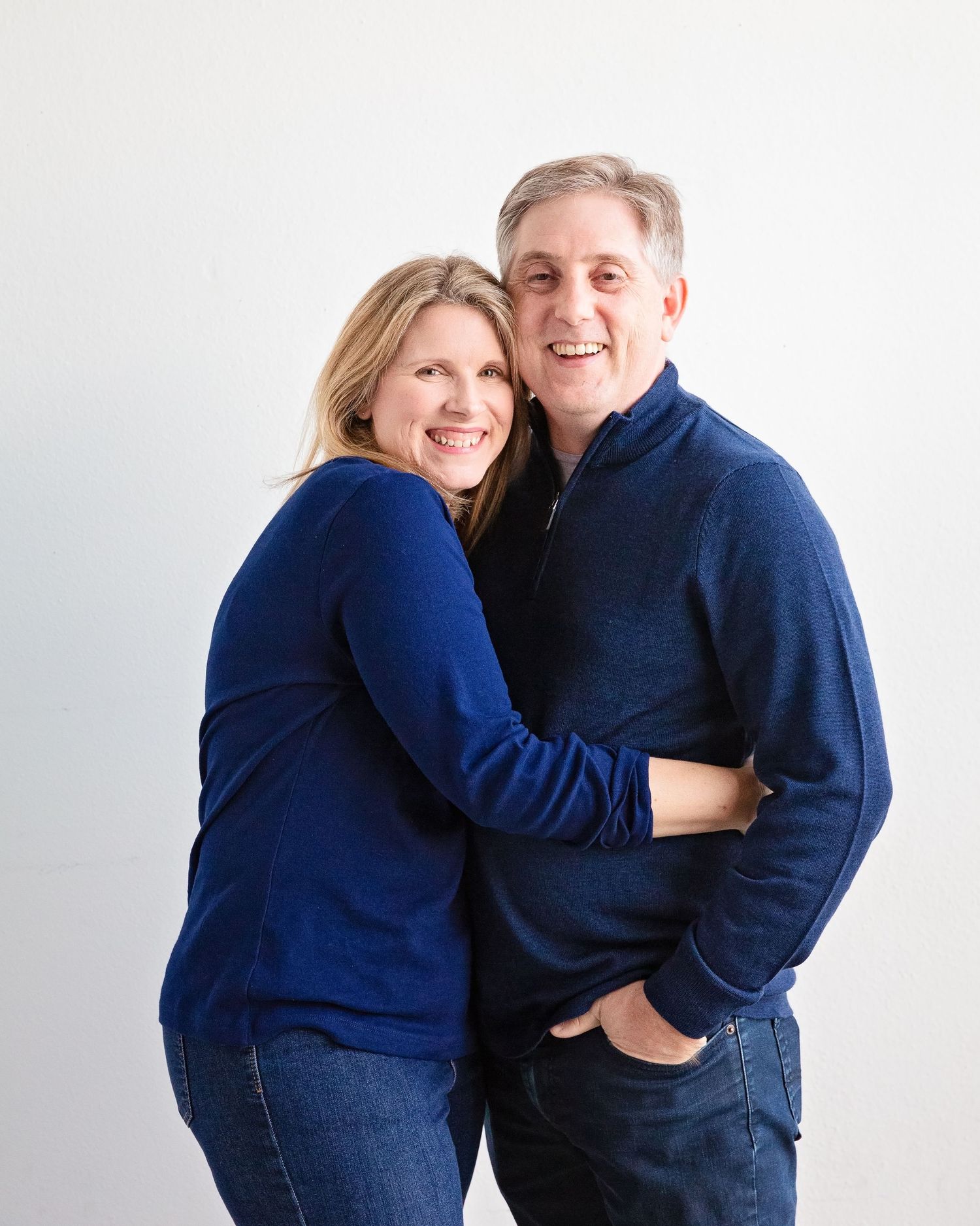 A couple in matching navy sweaters embraces against a white background in a casual portrait.