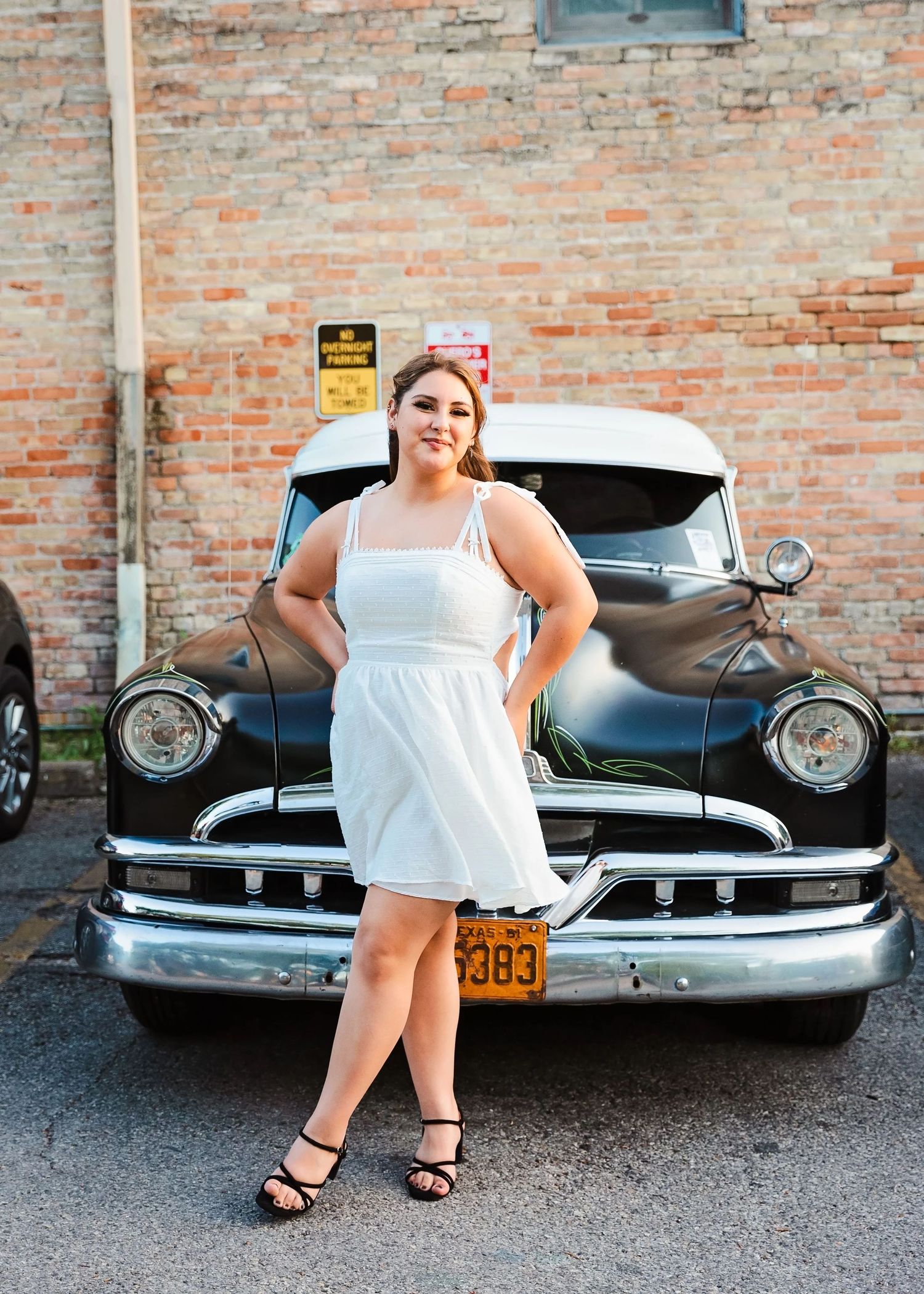 Woman in white sundress poses confidently in front of vintage black car against brick wall.