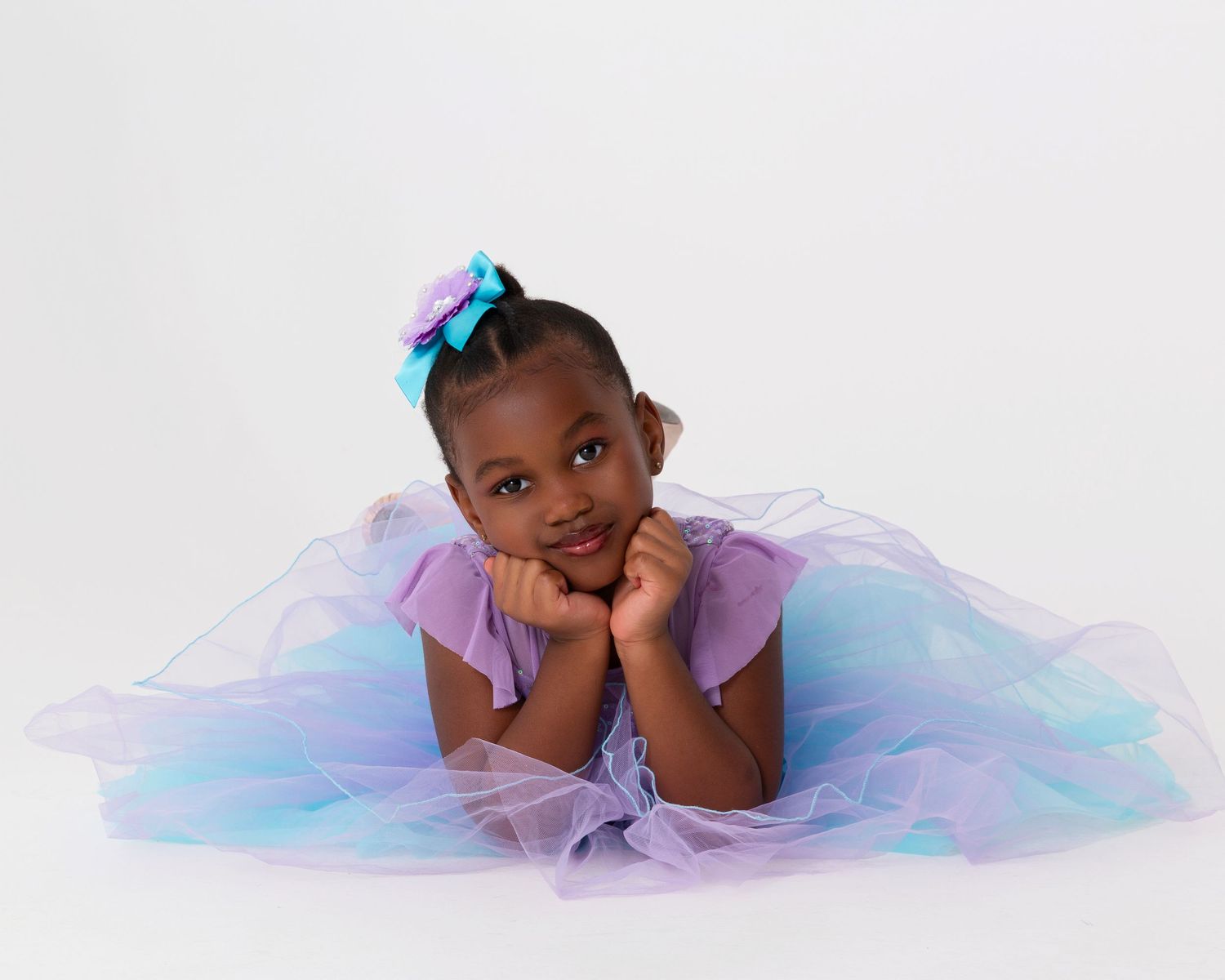 Young dancer in pastel blue and pink tulle dress lying gracefully on white background.