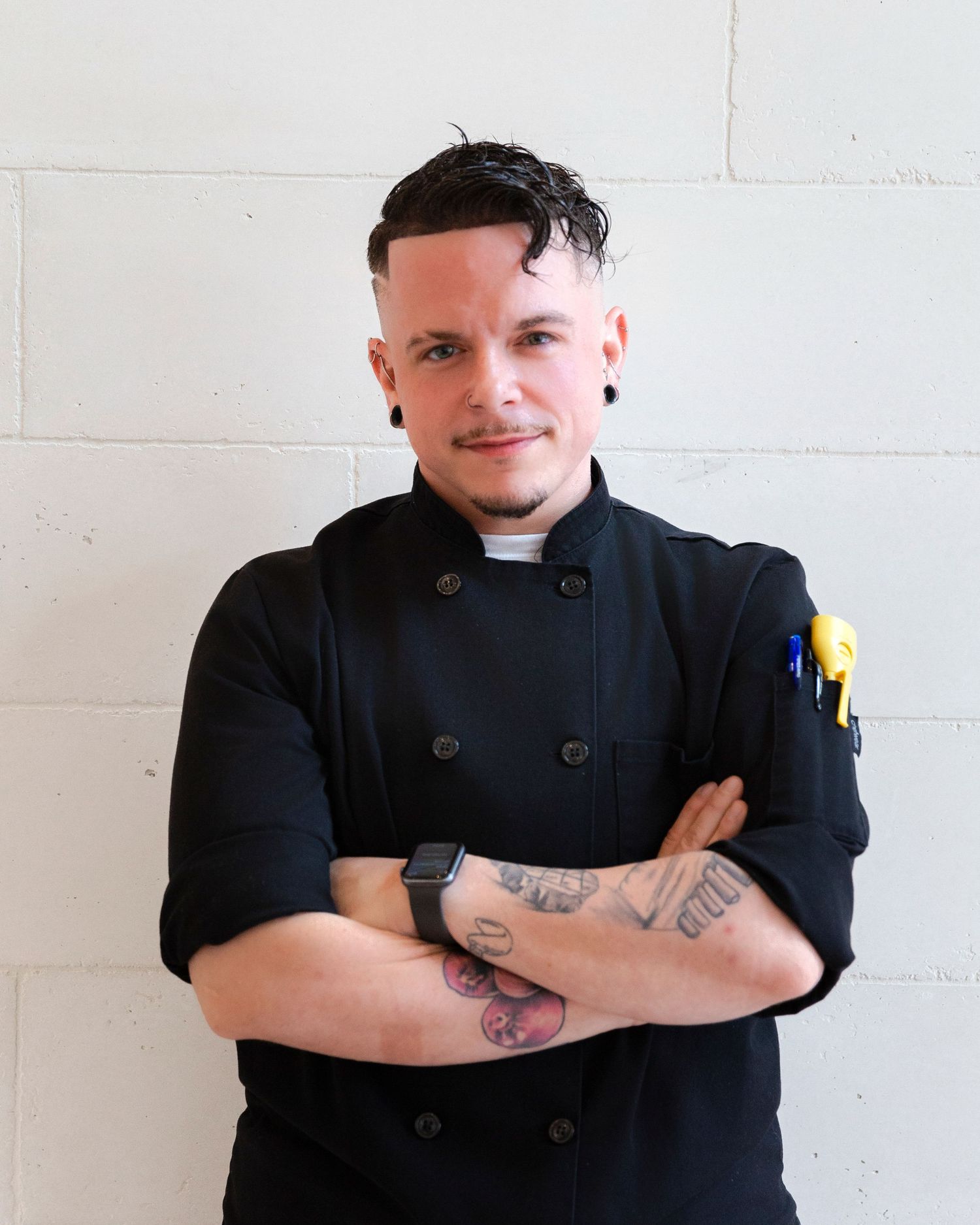 Chef in black uniform with tattoos poses against white wall.