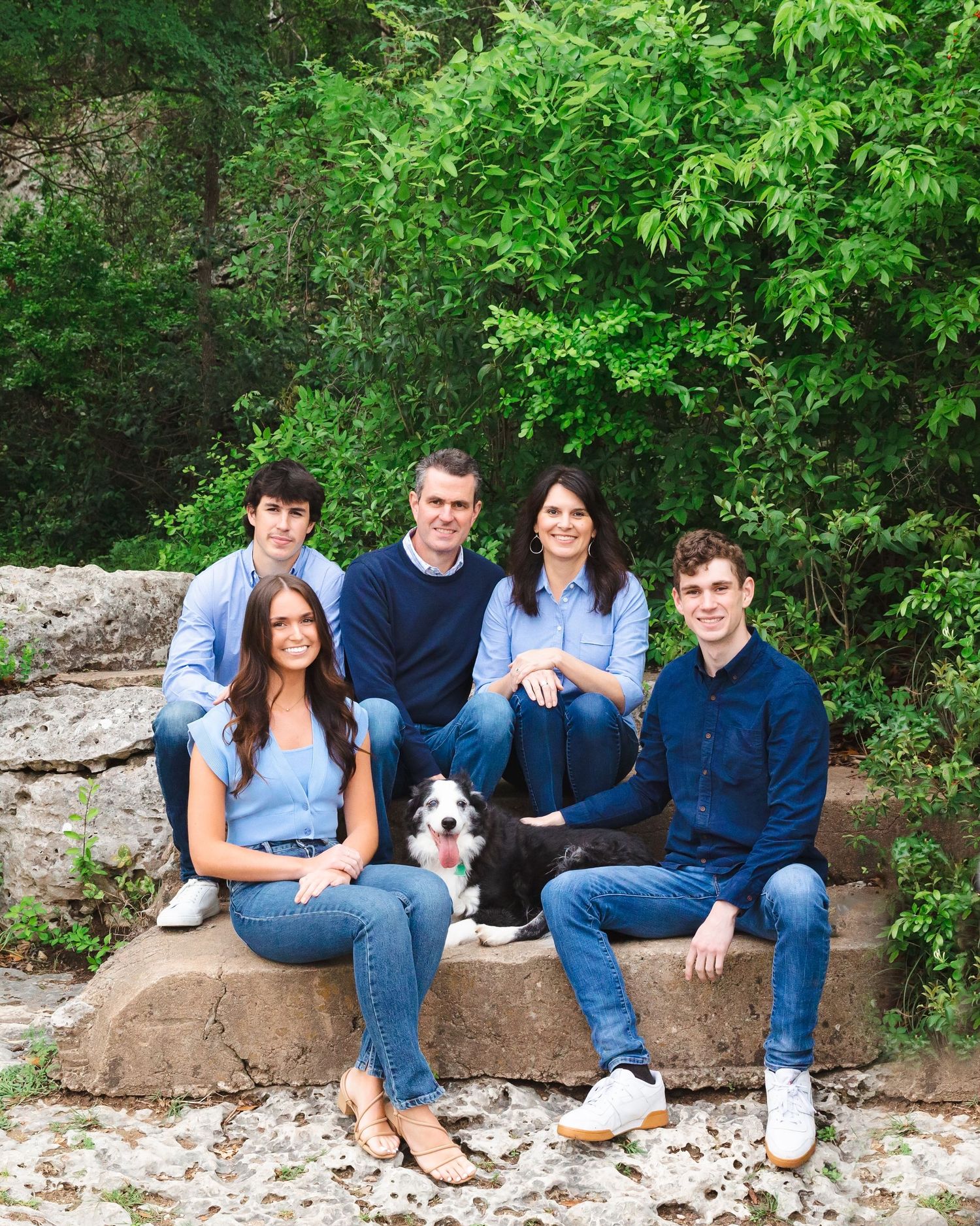 Family of five with dog sitting on large rock formation surrounded by green foliage.