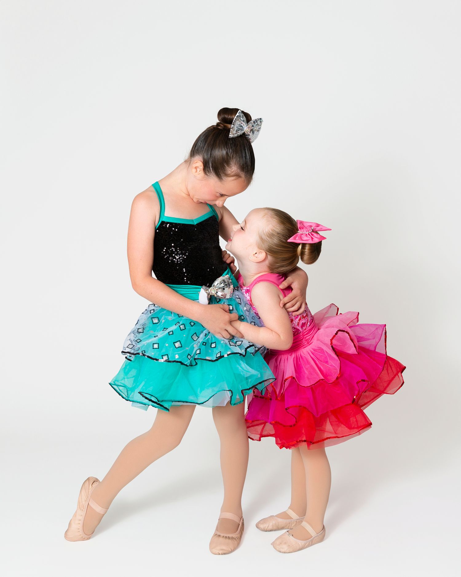 Two dancers in turquoise and pink tutus perform synchronized dance moves on white backdrop.