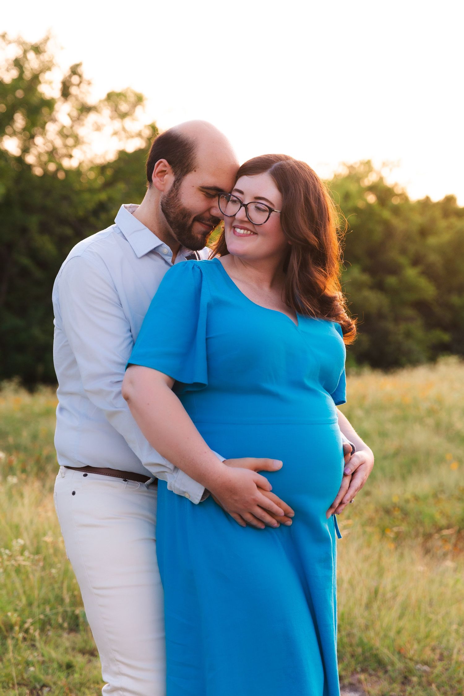 A couple poses in a field during their maternity session wearing coordinated blue and white outfits.