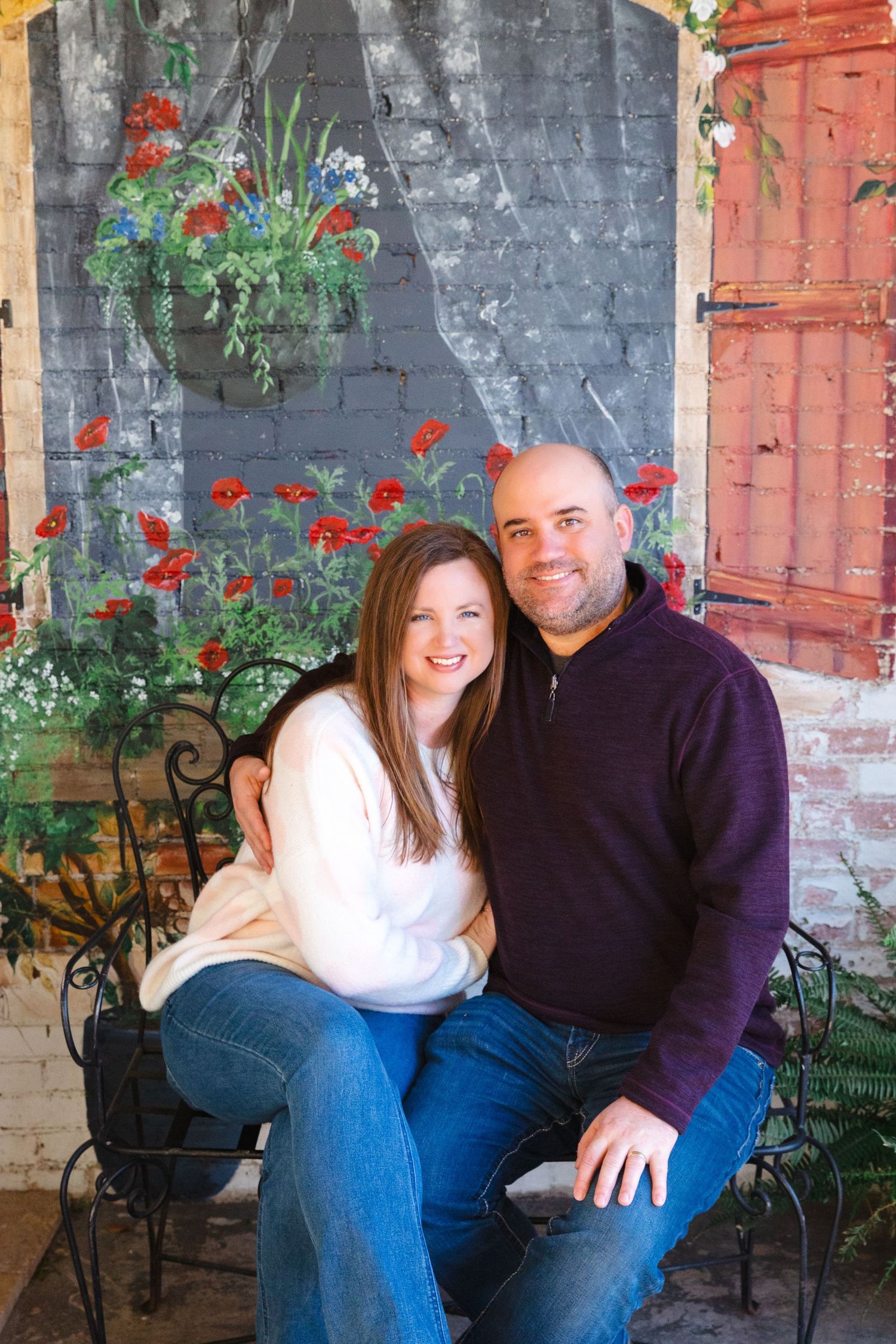 A couple poses together on a bench against a brick wall with red flowers and greenery.
