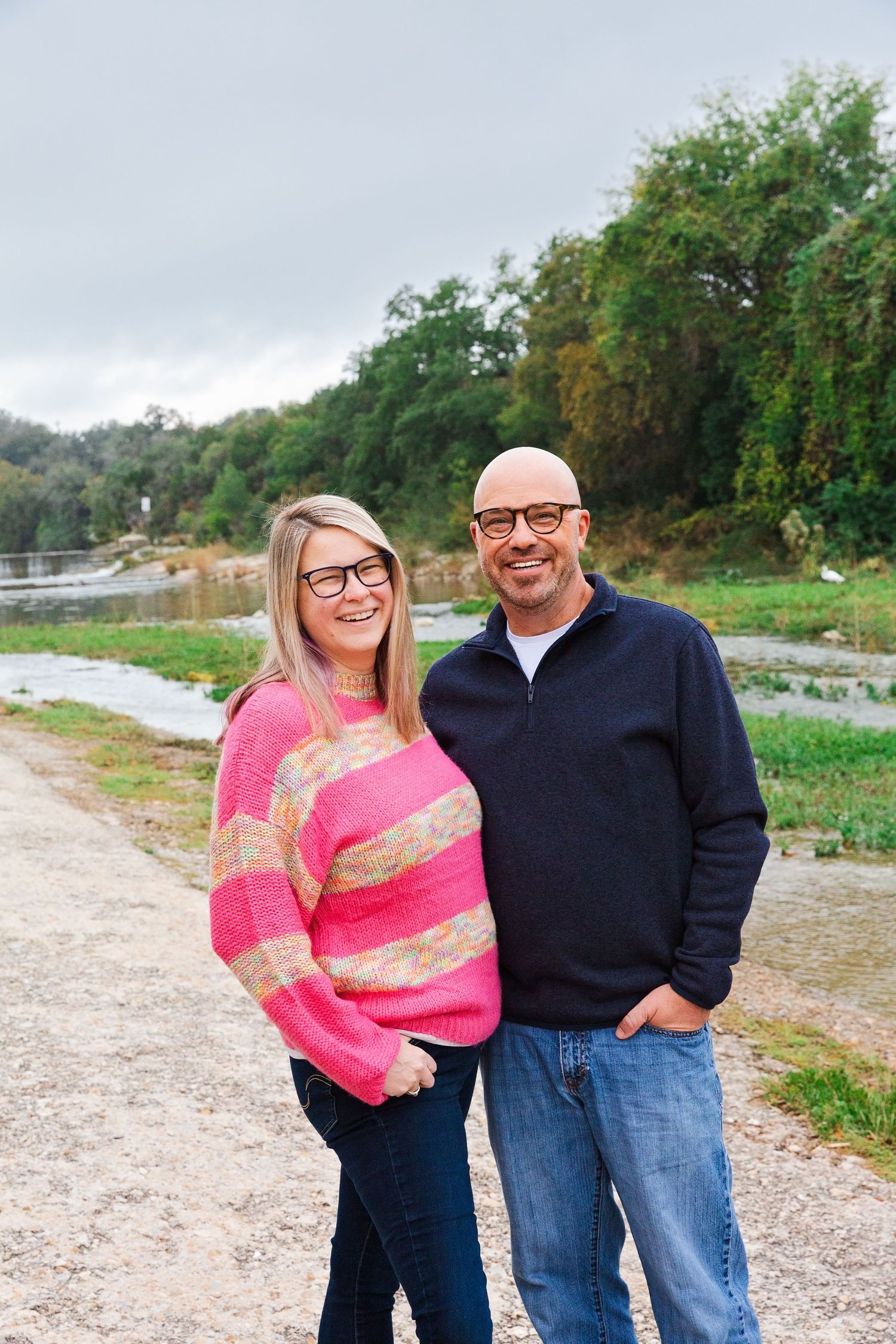 A couple poses together outdoors in casual attire with a pink sweater and black top against a natural backdrop.