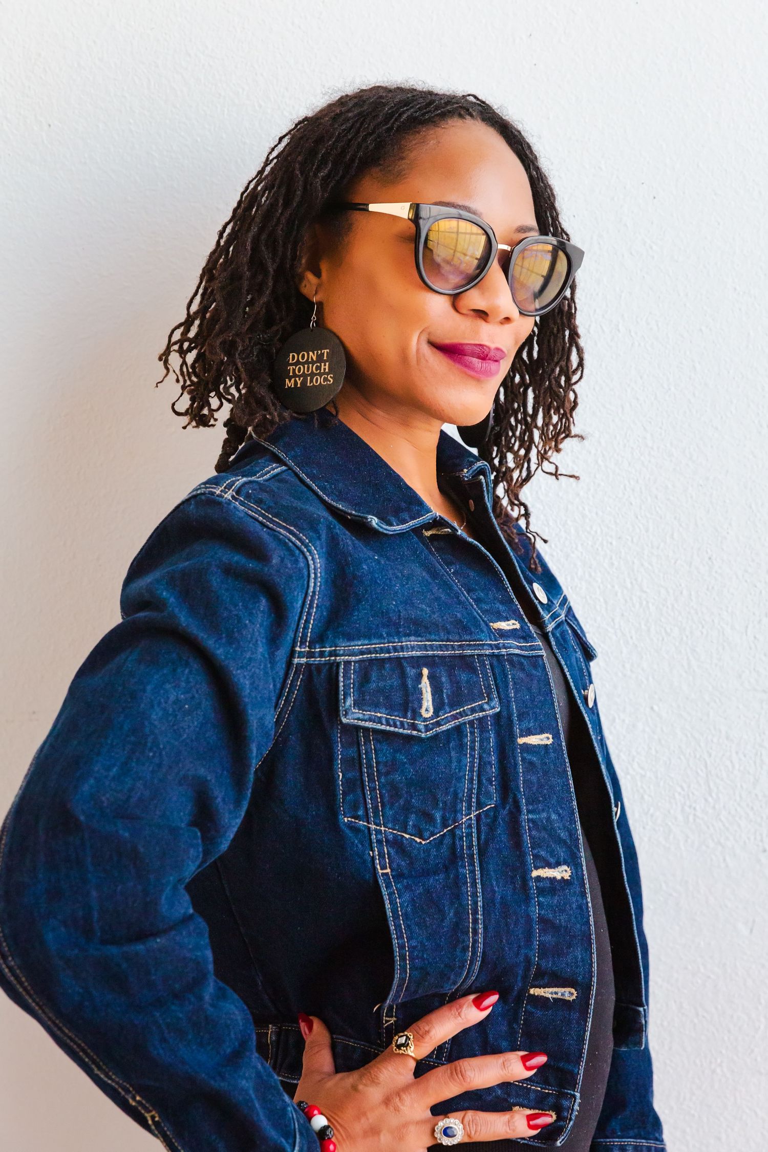 Stylish woman in denim jacket and glasses poses confidently against white wall.