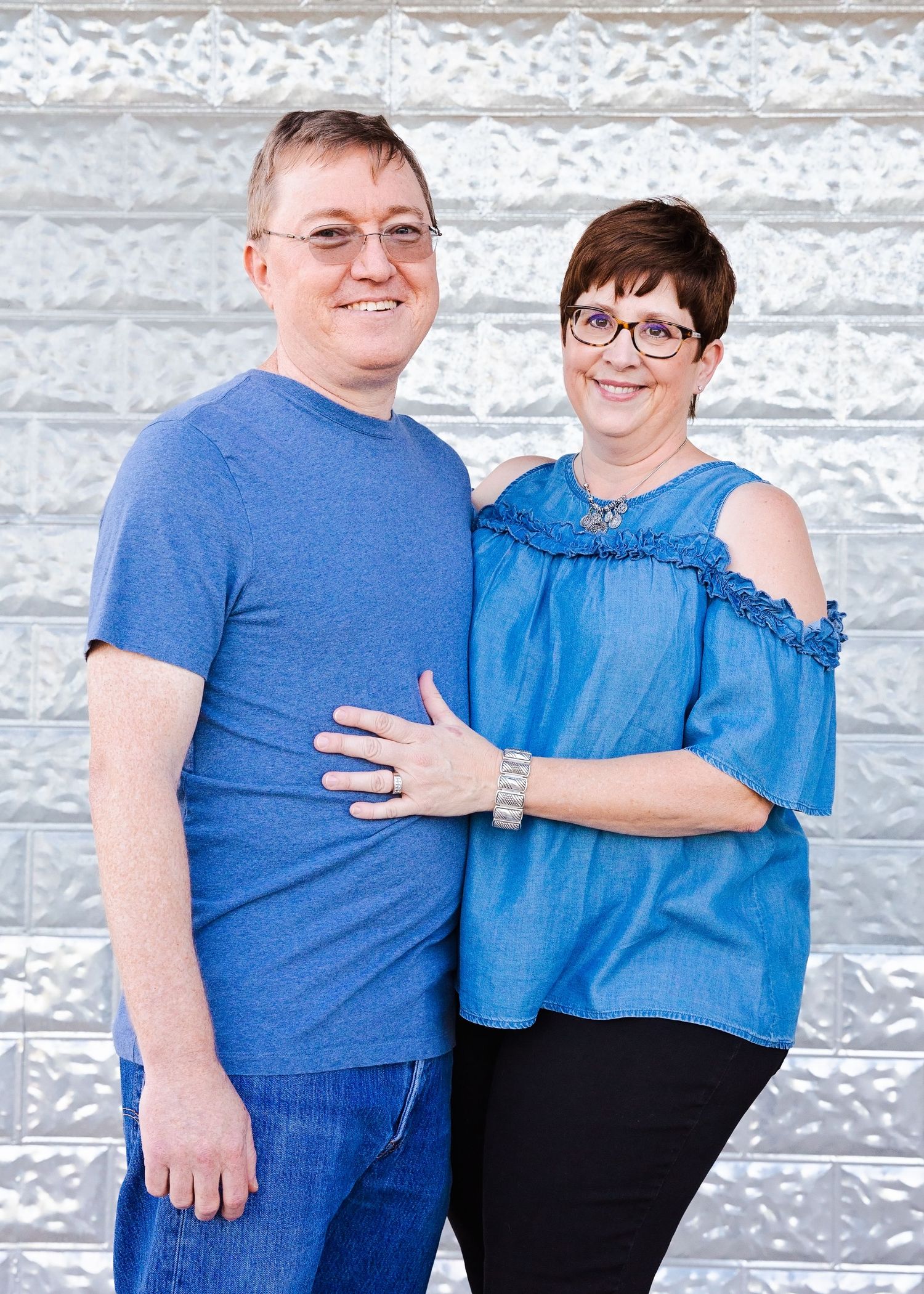 Two people in blue outfits smile together against white textured wall background.
