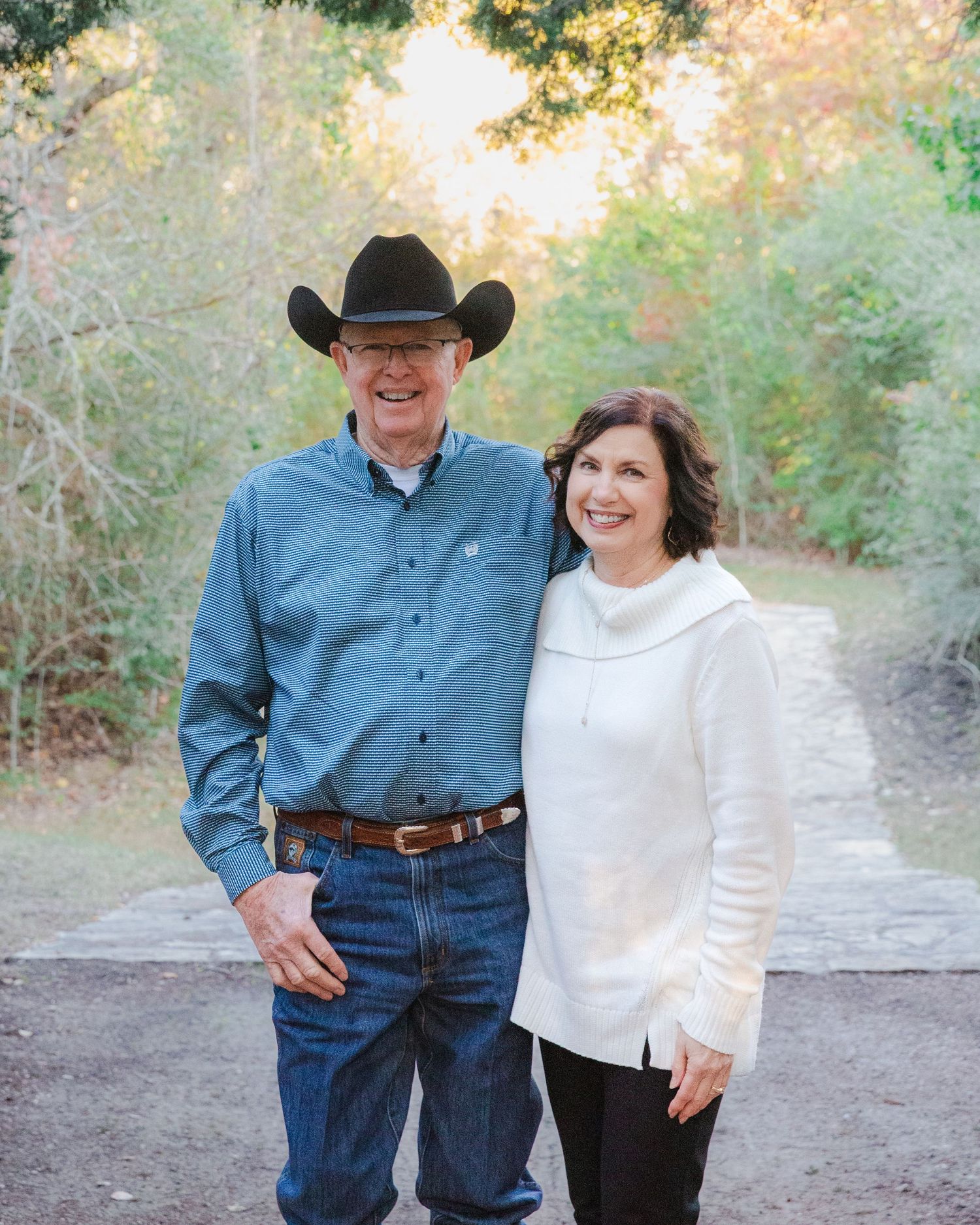 A couple in western attire poses together on a natural path during golden hour.