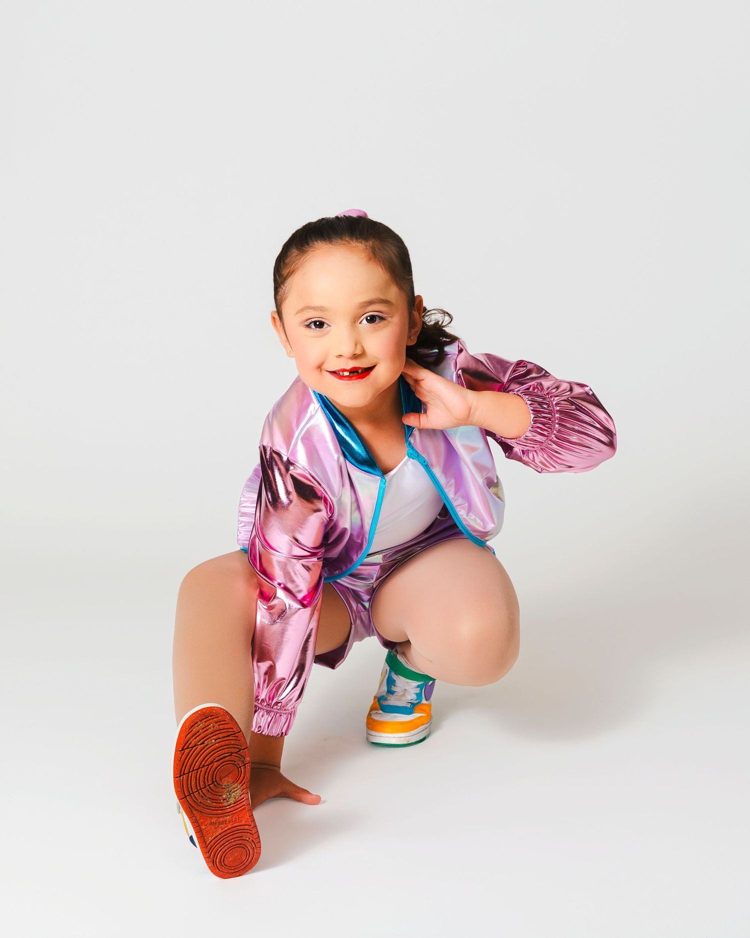 Dancer in pink metallic jacket and shorts strikes hip hop pose against white background.