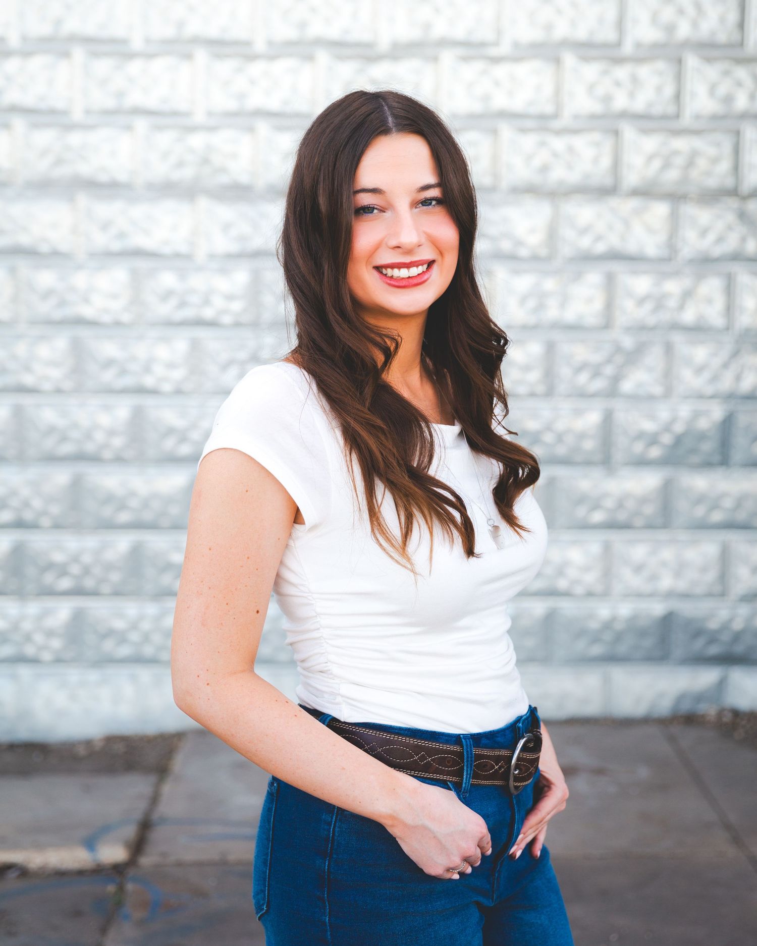 Senior portrait young Woman in white top and blue jeans poses casually against stone wall in outdoor portrait session.