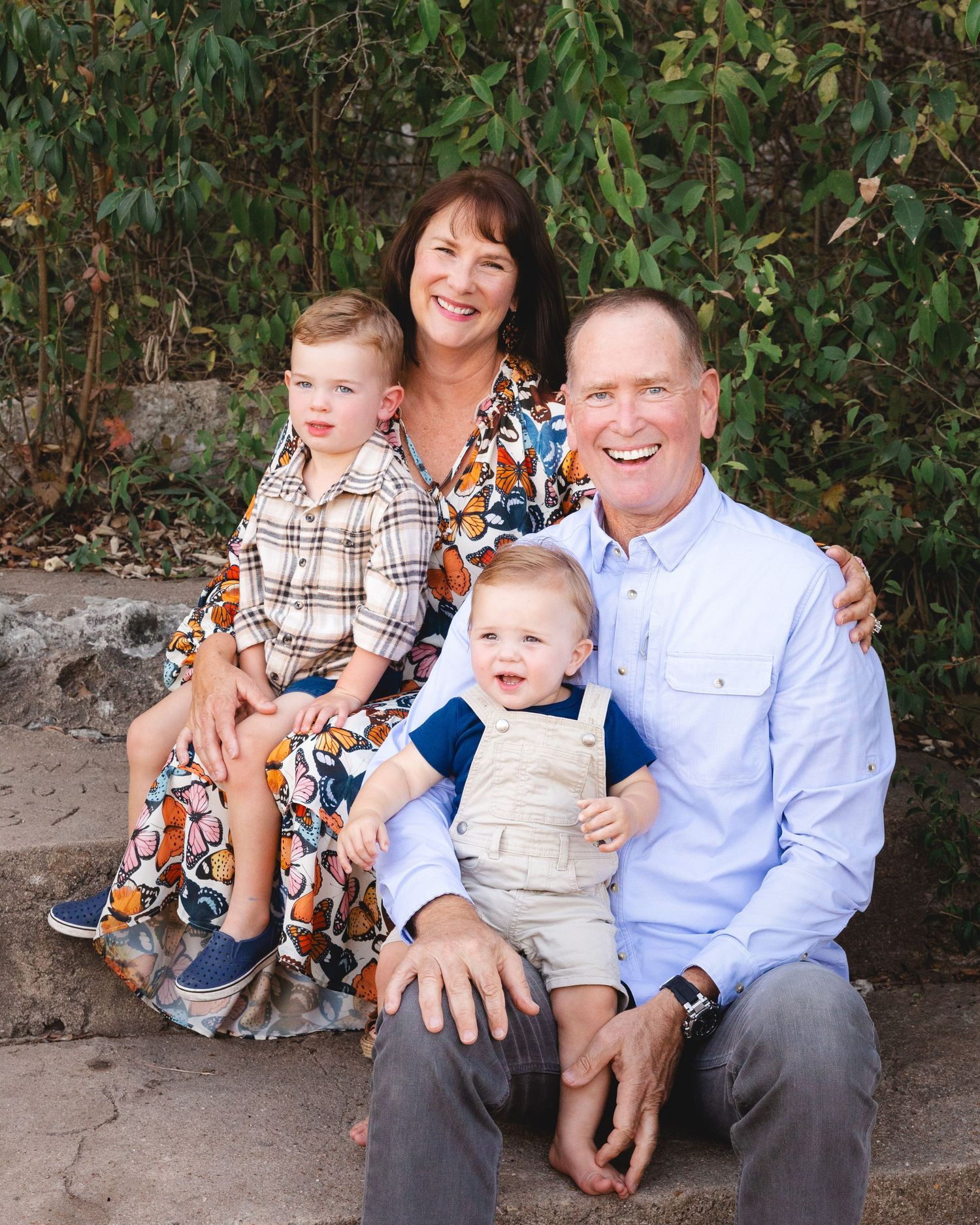 A casual outdoor family portrait session with people sitting on stone steps surrounded by greenery.
