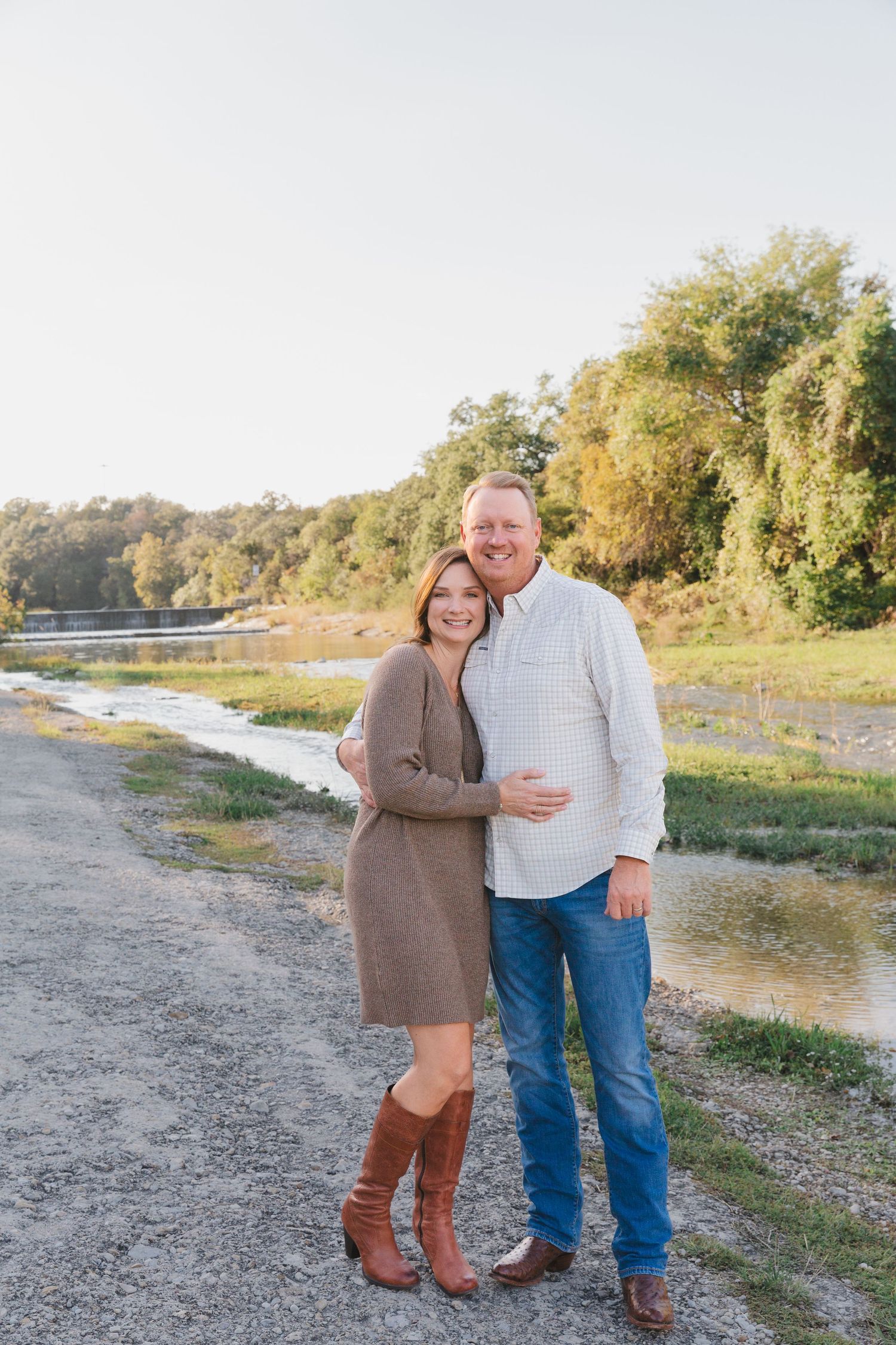 A couple poses together on a gravel path near a river during golden hour.