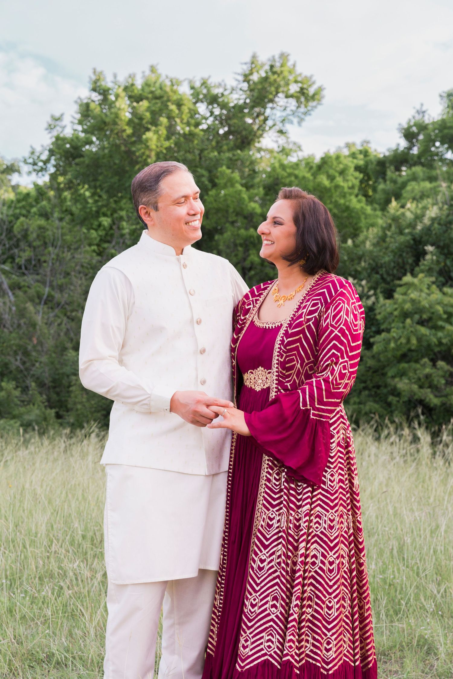 A couple in formal ethnic wear poses in a field, with white and burgundy patterned outfits.