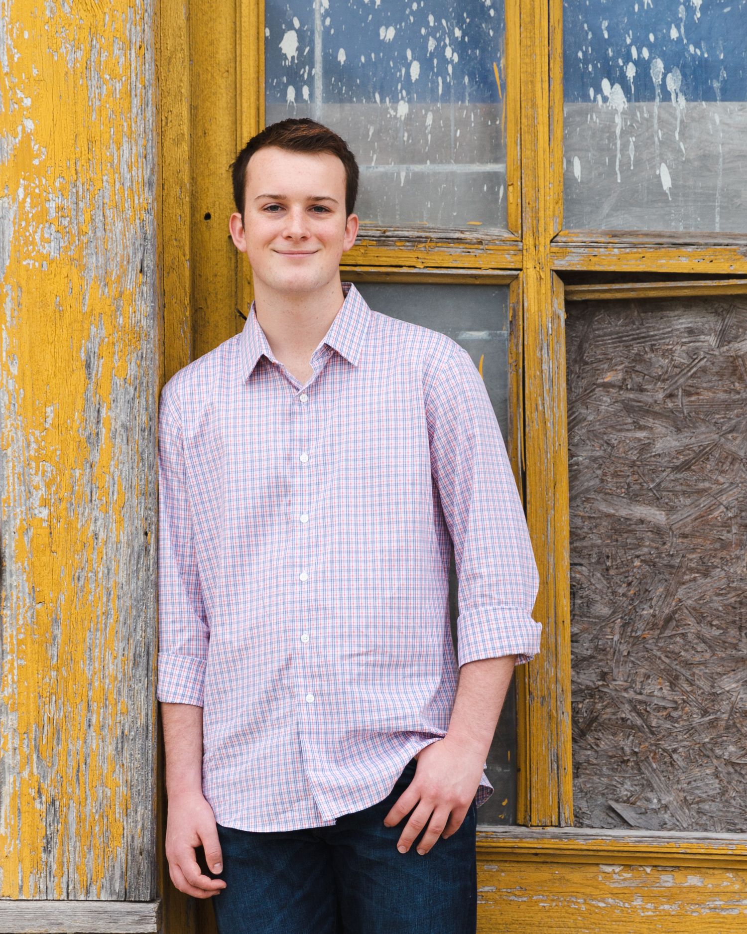 Person wearing a pink checkered shirt posing against a yellow and gray wall with windows. Senior boy portrait session in Round Rock, TX