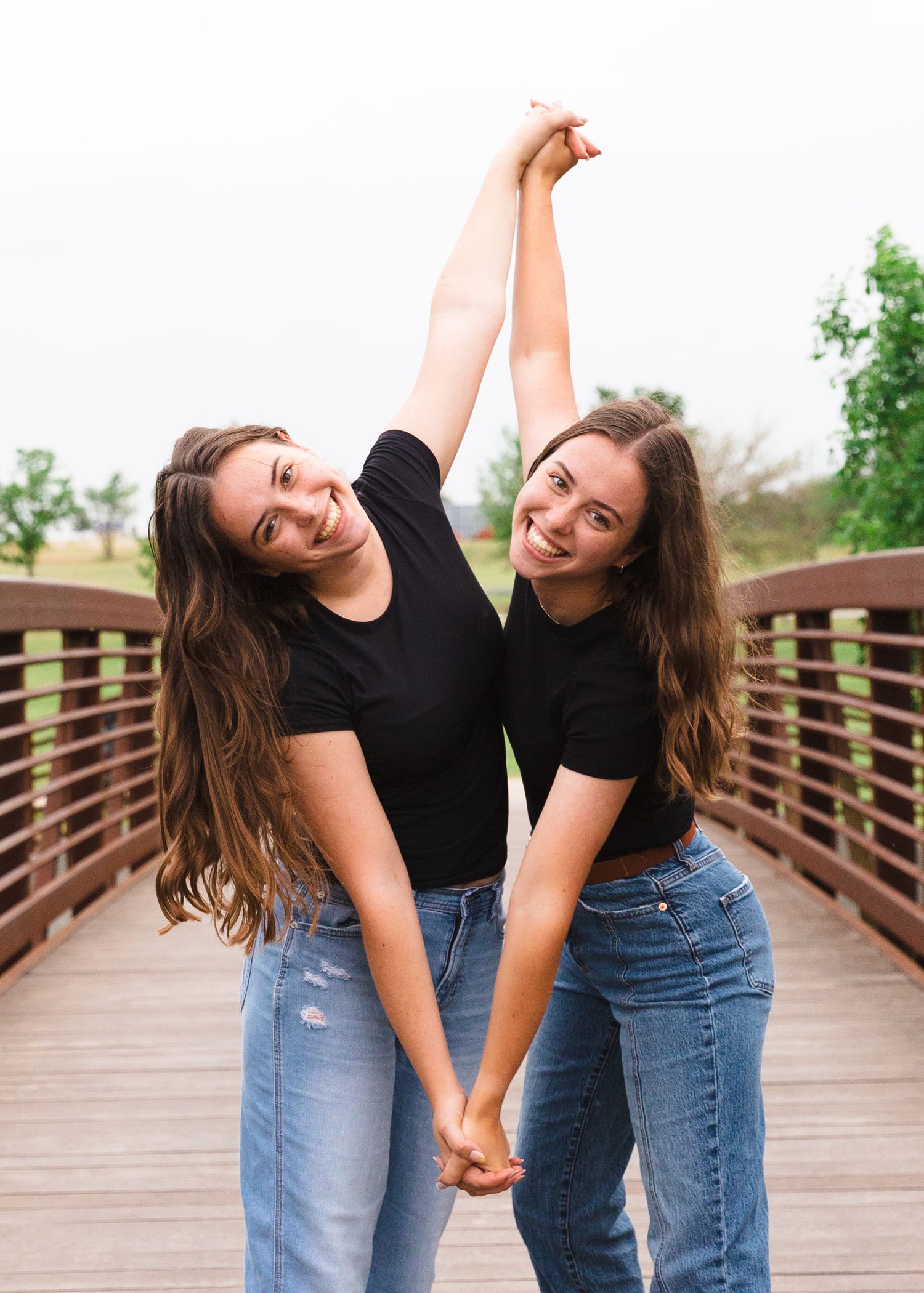 Two friends in black shirts and jeans hold hands while posing playfully on a wooden bridge.