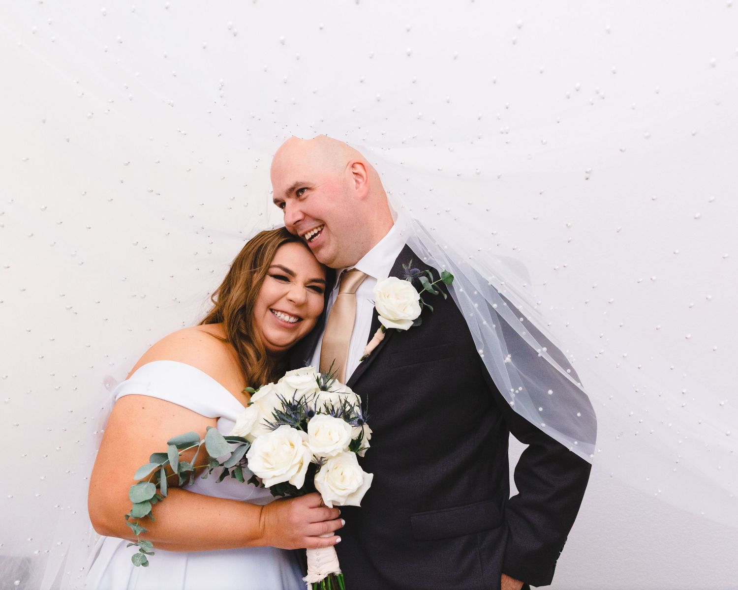 Newlywed couple sharing intimate moment with white roses against white wall.