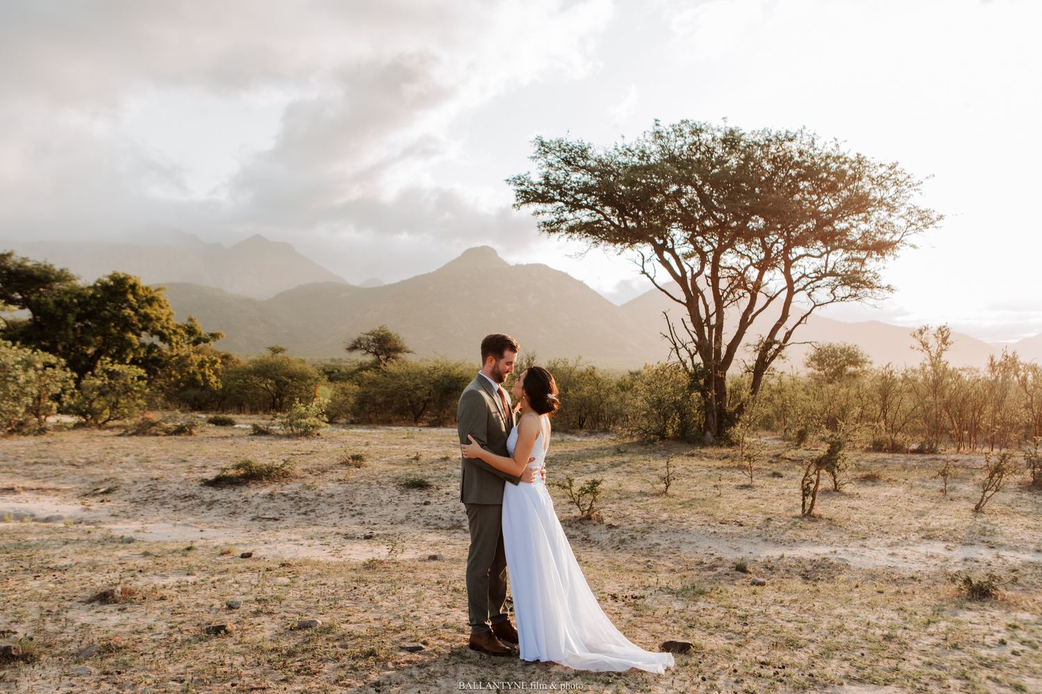 Wedding couple poses in desert landscape with mountains and acacia trees at sunset.
