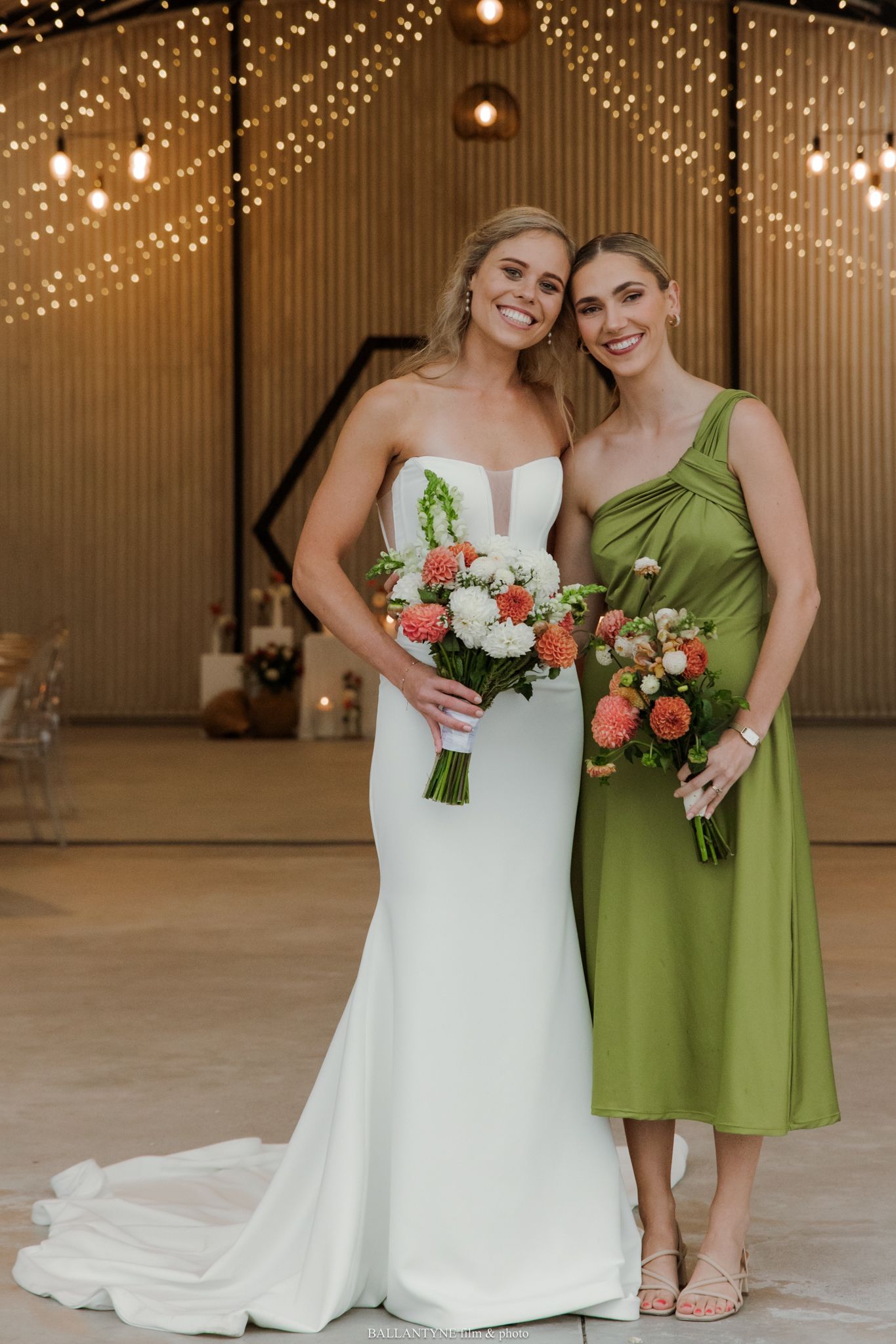 Bride in white gown and bridesmaid in green dress holding coral bouquets at elegant barn wedding venue.