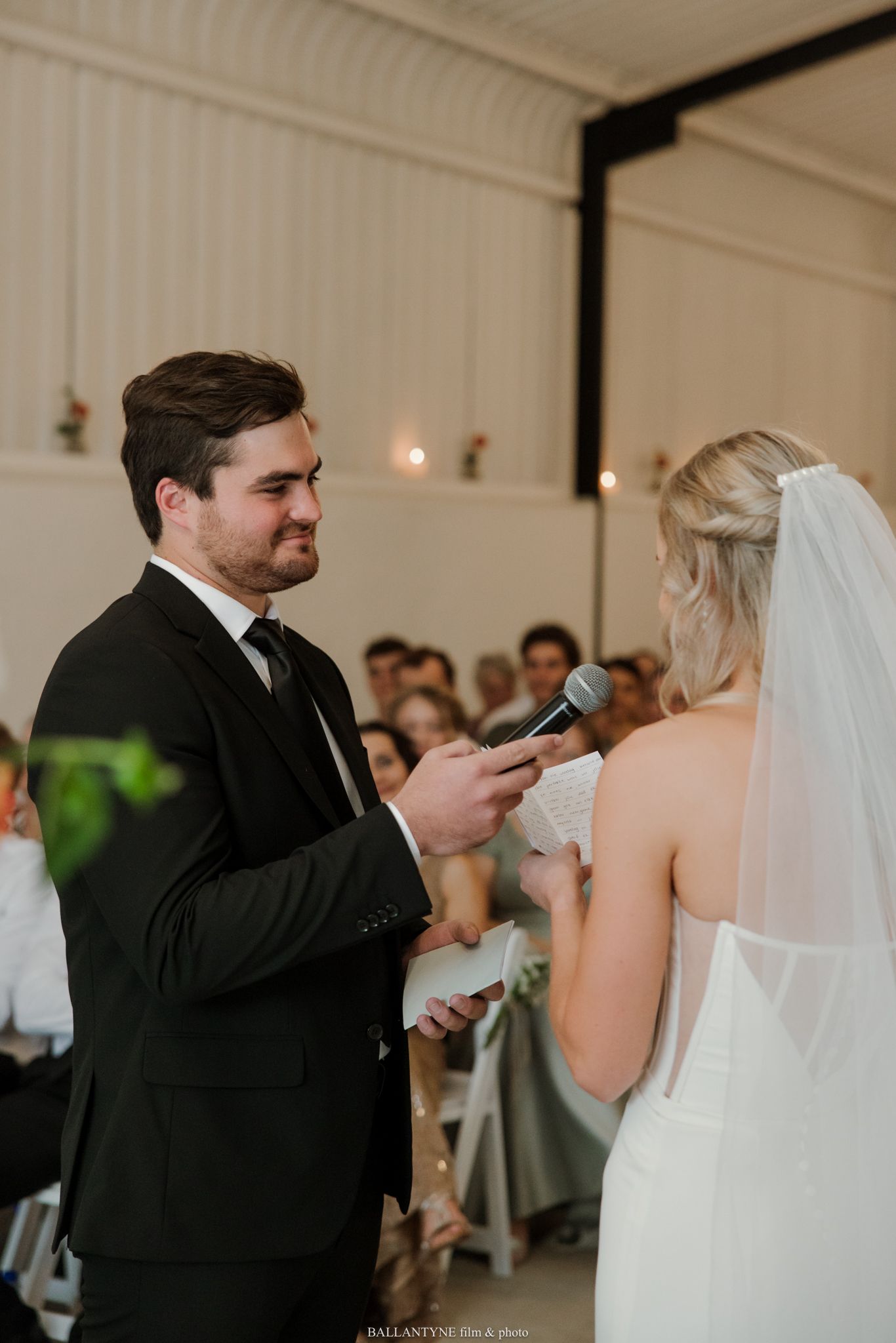 Groom reads wedding vows to bride at altar during indoor ceremony, holding microphone and paper.