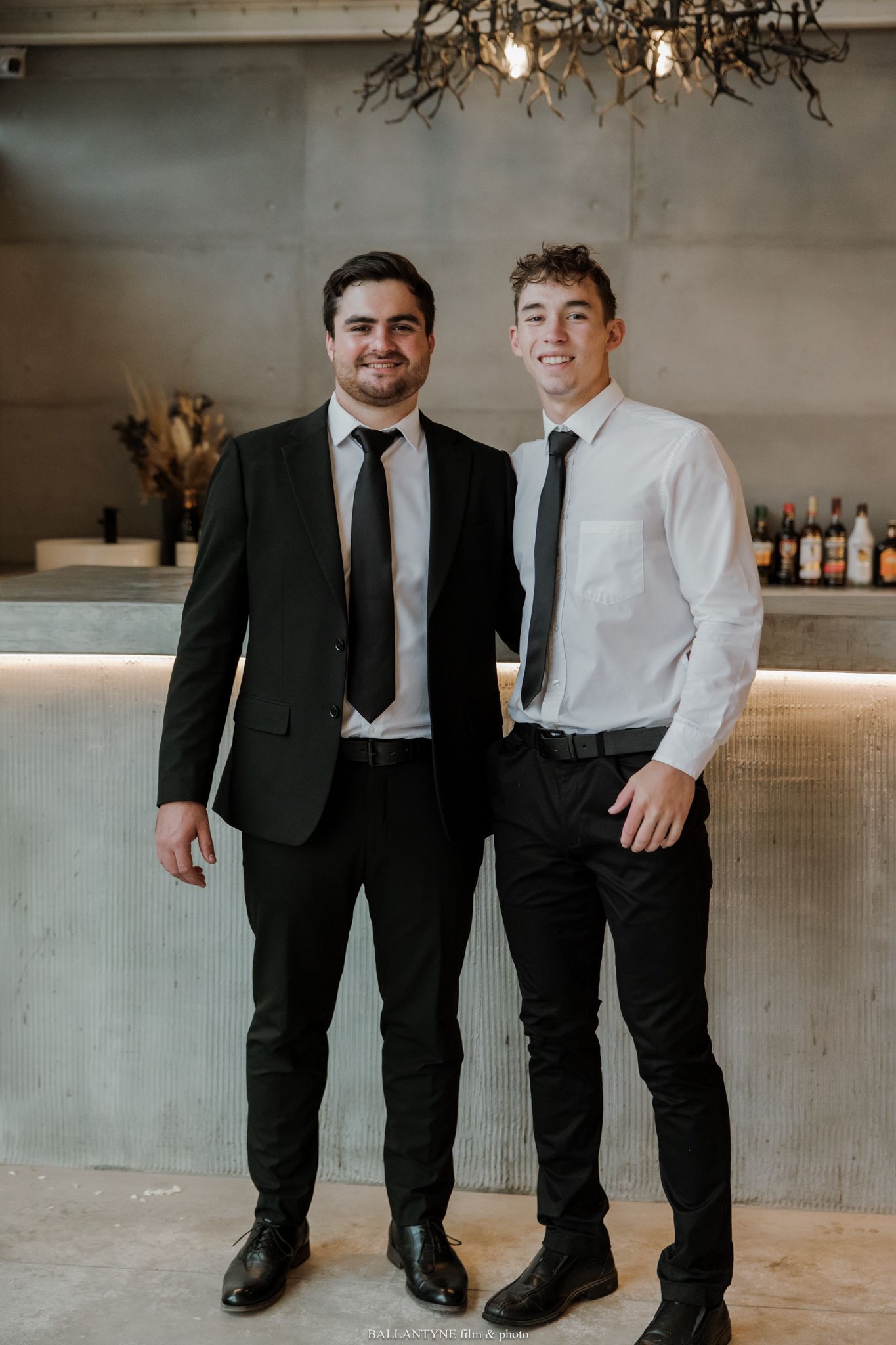 Two young men in black ties and formal attire smile together at an elegant bar venue.