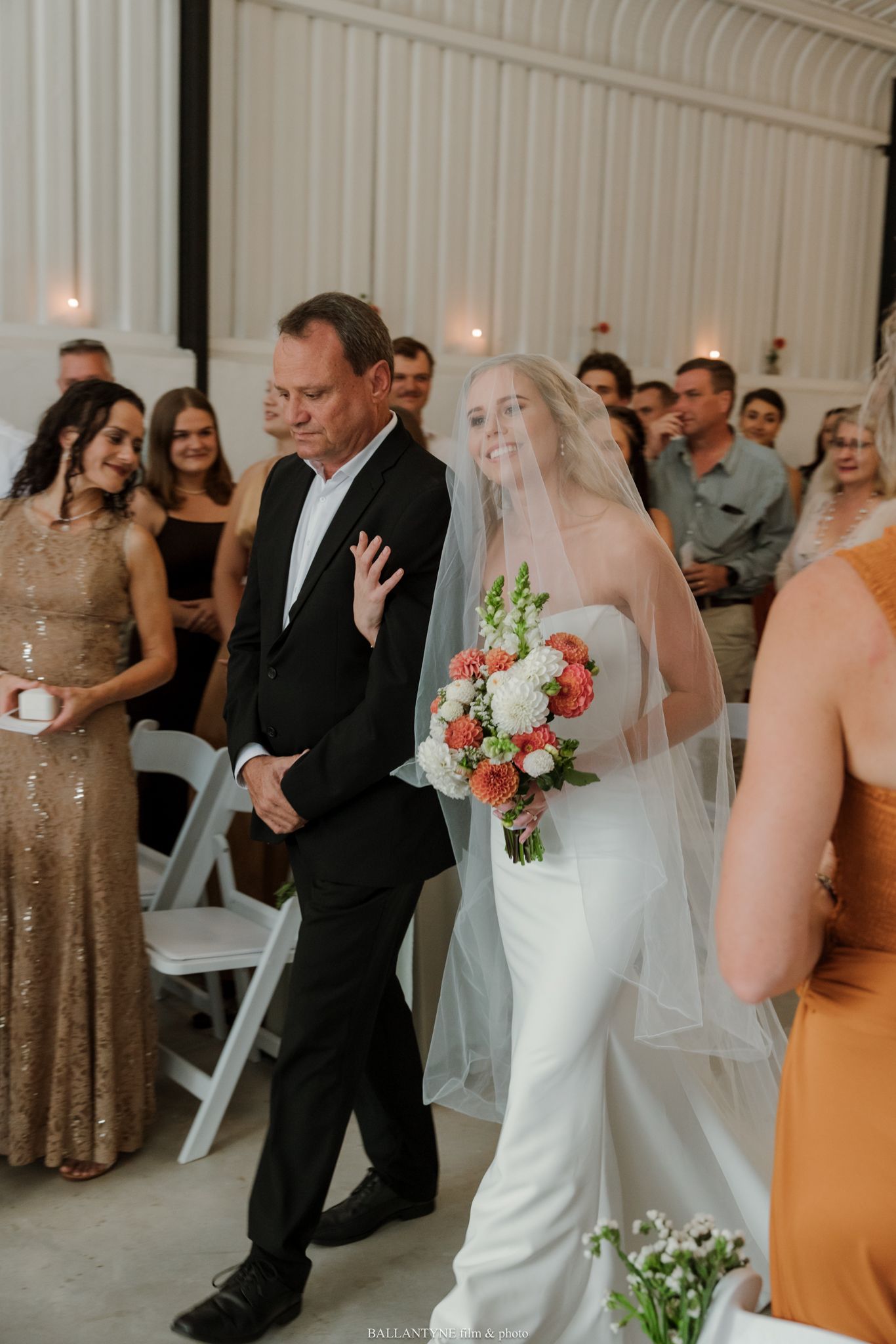 Bride walking down the aisle with her father, holding a coral and white bouquet, guests seated in white chairs.